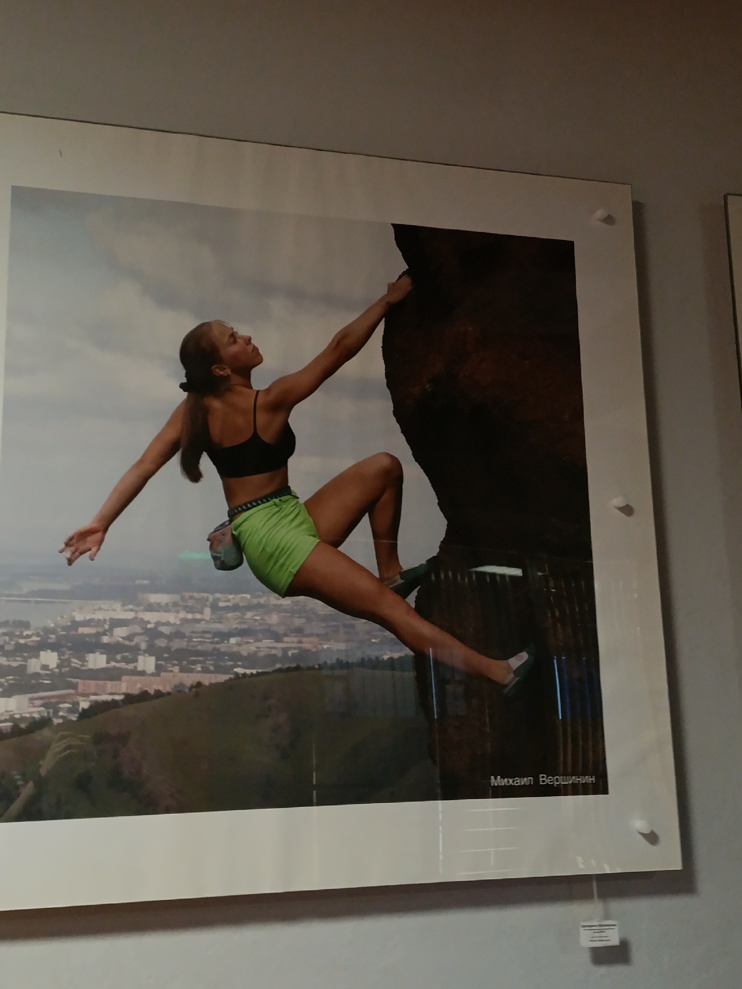 The image shows a framed photograph of a woman rock climbing, presumably outdoors. She is wearing a black sports bra and bright green shorts with a chalk bag attached to her waist. Her hair is pulled back into a ponytail. The woman is reaching up with her right hand to grip the rock face. Her left arm is extended outward for balance. She is also using her feet to secure herself on the rock.
In the background, there is a cityscape, which seems to be a town or city situated amidst hills and possibly near a body of water. The photo also has the photographer's name written on it, "Михаил Вершинин," which translates to Mikhail Vershinin.
The framed photograph is mounted on a wall, possibly in a gallery or exhibition space. The edges of the photograph and the mounting hardware are visible. The photograph is located in the city of Krasnoyarsk, Russia.