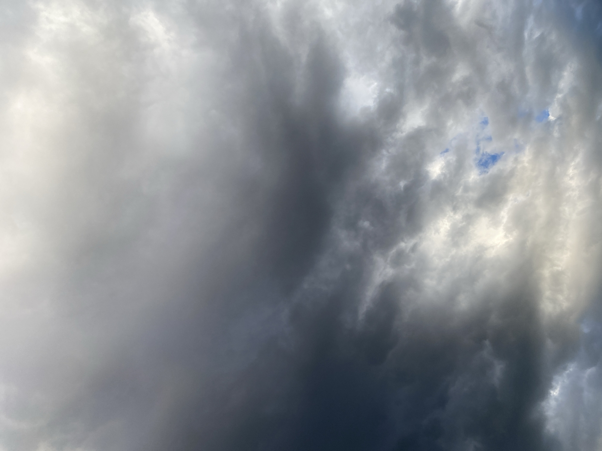 A wide-angle, low-angle shot captures a dramatic sky filled with dense, dark storm clouds dominating the left side of the frame. These dark clouds transition into lighter, white, and gray clouds towards the right, with glimpses of bright blue sky visible through breaks in the cloud cover. The lighting suggests either early morning or late afternoon, with sunlight diffused by the heavy cloud layer. The scene is devoid of any people, structures, or identifiable landmarks, presenting an abstract and elemental view of the sky above Байқоныс ауылдық округі, Kazakhstan.