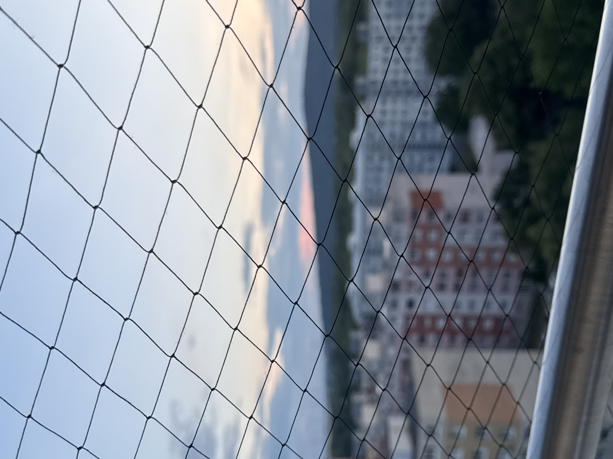The image shows a view through a black net with diamond-shaped openings. The net is in the foreground and is out of focus, creating a textured overlay on the scene behind it. The background features a blurred cityscape with buildings of varying heights and colors, including some with reddish-brown facades. Lush green trees are visible interspersed with the buildings, suggesting an urban setting with parkland or natural areas.
On the left side of the image, the sky is visible, with soft hues of blue, pink, and white, indicating either sunrise or sunset. The sky is partially obscured by a dark, blurry shape that could be a mountain or a large hill, separating the sky from the cityscape. The lighting suggests it is either early morning or late evening.
On the right side of the image, a metallic railing or bar runs vertically, also out of focus. The overall impression is a view from a balcony or window, possibly in a residential area, overlooking a city. There are no people or specific objects clearly identifiable due to the blurriness and the net obstruction. The location is given as Bratislava, Slovakia, which aligns with the presence of urban architecture and potentially the natural landscape features.