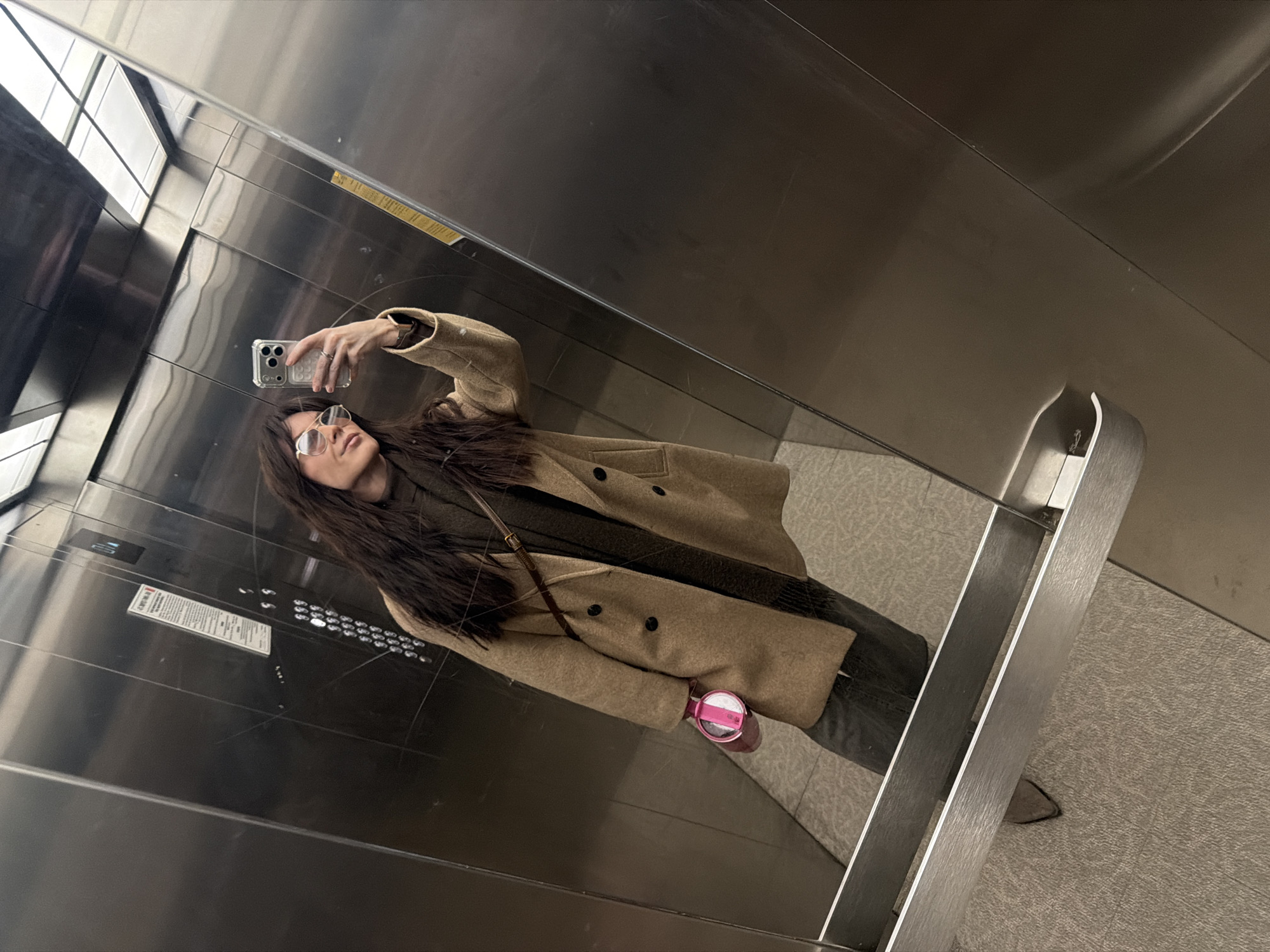 A lone female is depicted taking a selfie within an elevator. She has long, dark brown hair and is dressed in a light brown double-breasted coat over a dark green top, dark trousers, and dark, pointed-toe footwear. She wears gold-rimmed sunglasses and a ring on her right ring finger. A silver smartphone with a triple-lens camera array is held in her raised right hand, while a pink cylindrical thermos is held in her left. A dark brown bag strap crosses her upper body. The elevator's interior walls and doors are constructed of polished dark metallic material, reflecting the scene. A control panel with multiple circular buttons is on the left wall. An informational sticker, primarily in Russian, is affixed to the left wall, indicating the location as TRC "Iceberg" at Petropavlovskaya Street, 59, Perm, Russia. The floor is covered with a light-colored, patterned carpet.