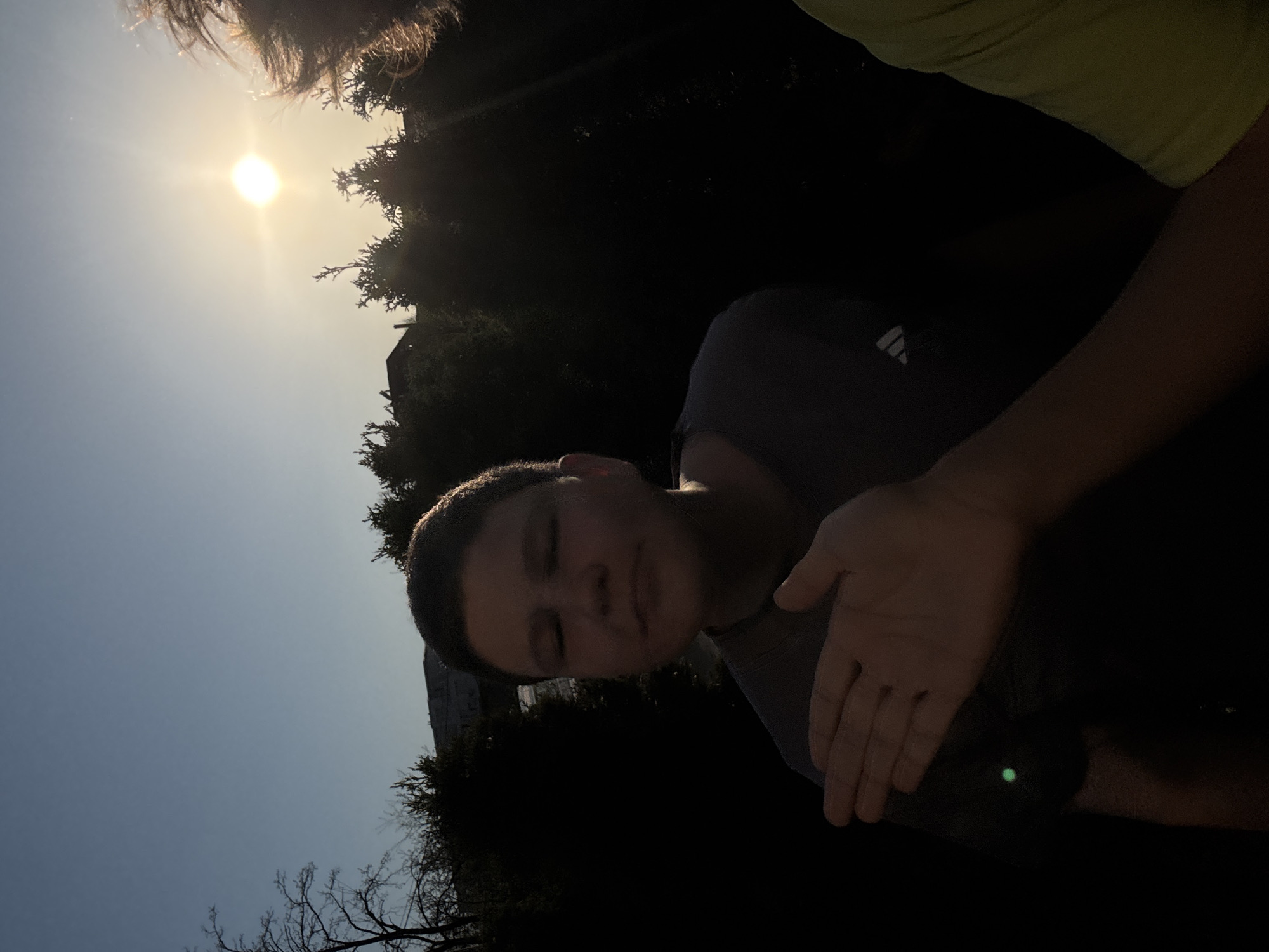The image shows a person with short, dark hair and closed eyes, wearing a gray t-shirt with an Adidas logo. Another person's arm, wearing a light green shirt, is partially visible in the upper right corner. The scene is outdoors, with a bright sun in the upper left and trees creating dark shadows. A building is visible in the background, suggesting an urban setting. Although the lighting is poor, based on the architecture, foliage, and sunlight, the location is potentially in a park in Sofia, Bulgaria, although more visual information would be required for verification.