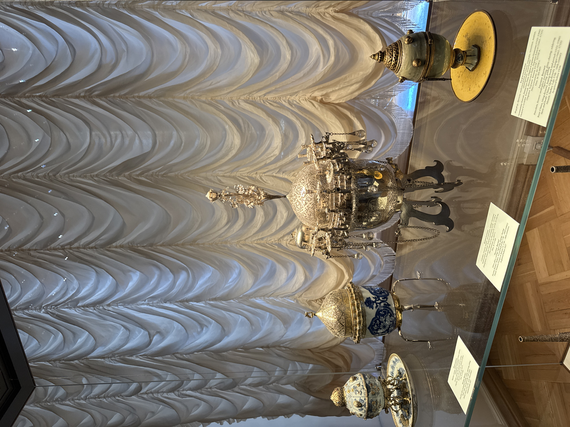 The image depicts a display of decorative artifacts, possibly in a museum setting, situated in Saint Petersburg, Russia. The background is dominated by a large, off-white curtain with elaborate draping and pleating, creating a theatrical backdrop. In the foreground, a glass display case showcases several ornate objects.
To the left, a small, blue and white porcelain object with a golden lid rests on a white plate. Adjacent to it is a larger, similar porcelain piece, also with a blue and white design, adorned with gold accents and topped with a golden dome featuring intricate detailing and a small crescent moon finial. This piece is supported by three slender, ornate legs.
In the center, a substantial, highly decorated silver or silver-plated vessel takes pride of place. It features a domed lid with elaborate filigree work and is adorned with numerous hanging crystal-like elements. This central piece appears to be a decorative urn or censer, supported by a multi-legged ornate base.
To the right, a small, greenish-blue egg-shaped ceramic object with golden accents sits on a bright yellow oval plate. This object has a layered, somewhat conical lid and appears to be a decorative box or censer.
Two small, white descriptive plaques are visible on the glass surface of the display case, providing information about the artifacts. The flooring beneath the display case is made of polished light-colored wood, laid in a parquet pattern. The lighting in the scene appears to be soft and ambient, highlighting the reflective surfaces of the metallic objects and the sheen of the curtains. No people are visible in the frame.