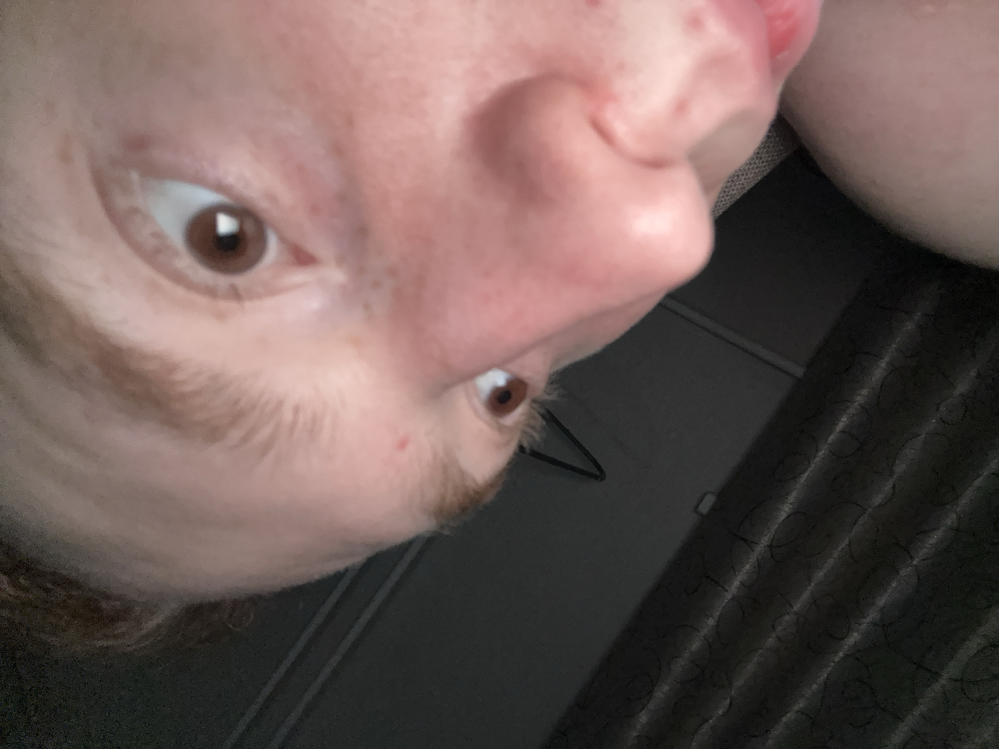 A close-up selfie oriented at a 45 degree angle, showing a person's face, including brown eyes, nose, and reddish facial hair. The background is dark, with a patterned surface and a glimpse of a wall and hanging object. There is no definitive information to confirm that the picture was taken in the city of Chaikovsky, Russia.