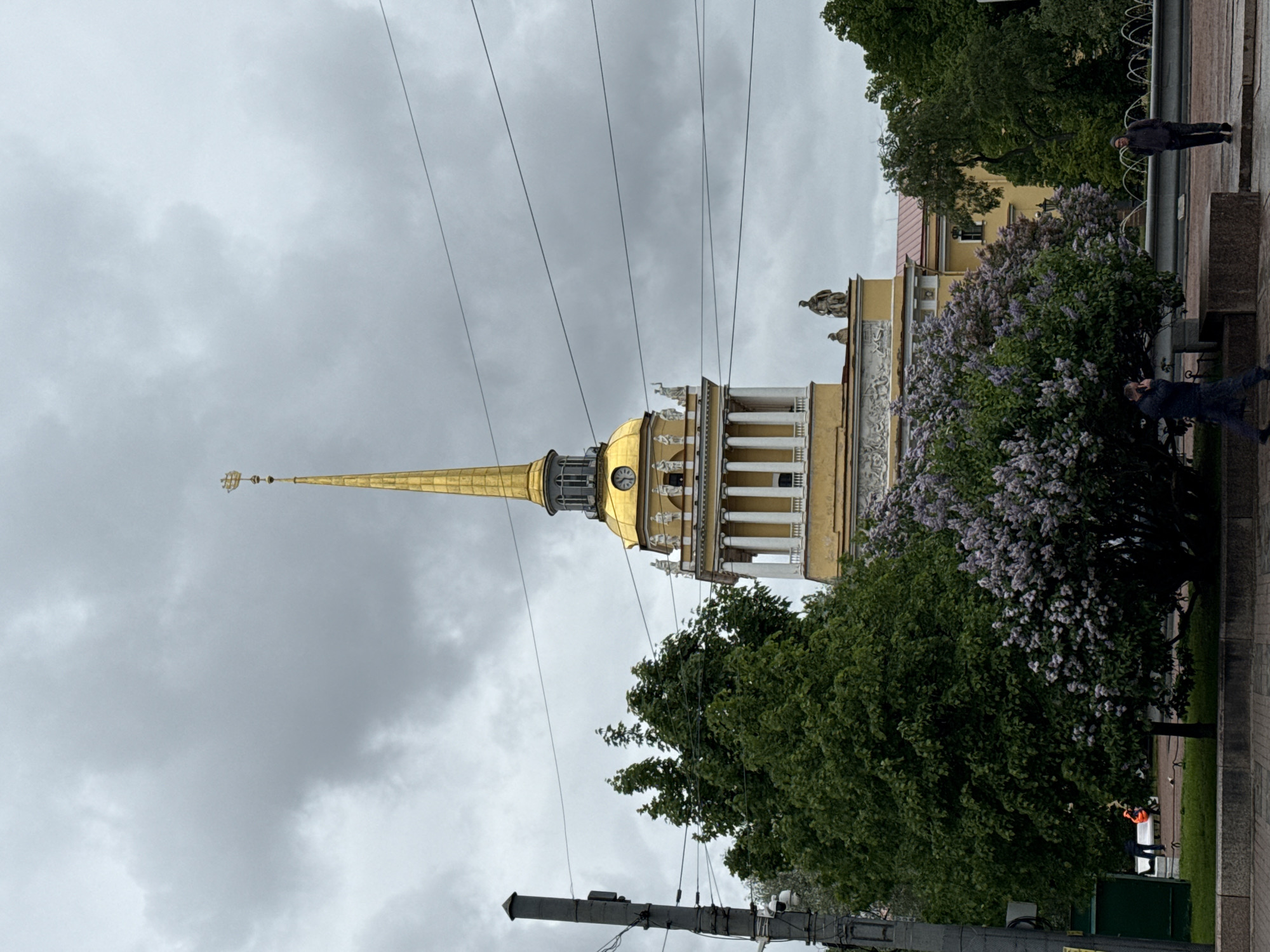 The photograph shows the spire of the Admiralty building in Saint Petersburg, Russia, framed by trees and lilac bushes against an overcast sky. The golden spire, a prominent landmark, rises from the building's columned structure with a visible clock face. Several power lines intersect the view of the spire. Two individuals stand on a low wall to the right of the frame. Another individual, who seems to be wearing an orange vest, appears at the bottom edge of the photograph. A lamppost with visible wiring is positioned at the bottom left. The presence of blooming lilac bushes suggests a spring or early summer setting.