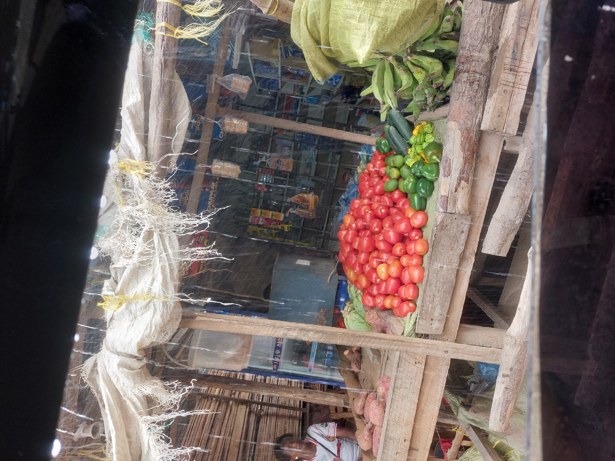 The image captures a bustling scene at a roadside market stall in Dar es-Salaam, Tanzania. The stall is constructed from rough wooden planks and is covered with white sacks and clear plastic sheeting. Inside, a variety of fresh produce is on display, including a large pile of ripe red tomatoes, green bell peppers, cucumbers, green bananas, and sweet potatoes. In the background, there are shelves stocked with packaged food items and soft drinks, suggesting the stall also functions as a small convenience store. A young woman wearing a white and red shirt is visible tending to the stall. The surrounding environment suggests a busy urban area.