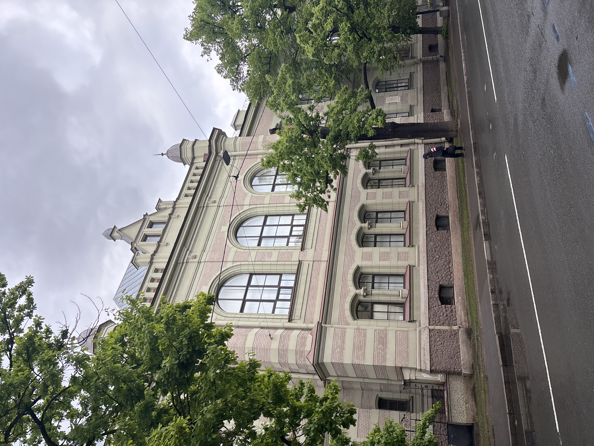 The image shows a building on a street in Saint Petersburg, Russia. The building has a light-colored facade with pink brick detailing and arched windows. Trees obscure parts of the building. A person is standing on the sidewalk next to the building. The street is wet and has a white line marking the lanes. The sky is overcast.