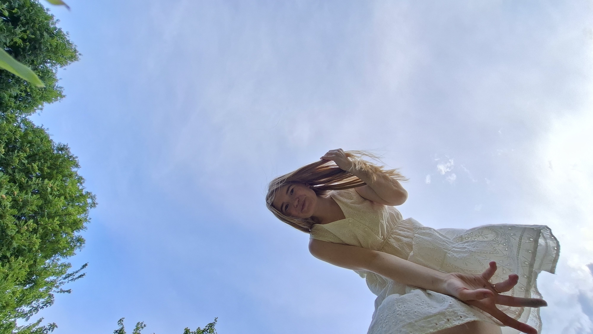 A low-angle shot captures a young woman against a backdrop of a blue sky and greenery. She is wearing a white dress. Her blonde-brown hair is partially obscuring her face. She is smiling and looking down at the camera, her right hand is raised towards the viewer, with her fingers spread apart. A few trees with green foliage are visible in the lower left and upper left corners. The background consists of a clear blue sky with some wispy clouds. There is no clear indication of this being in the city of Большой Букор, Russia.