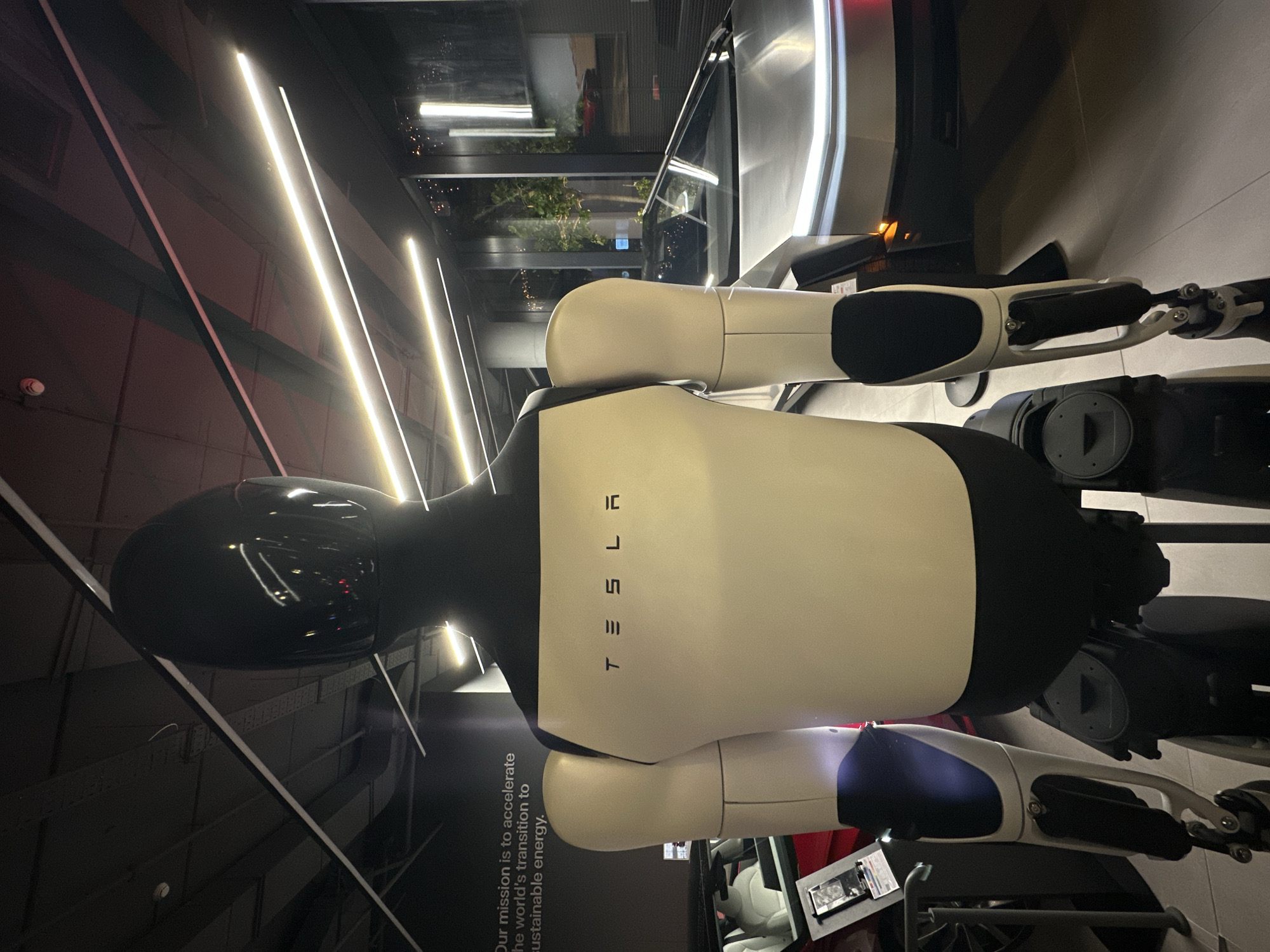 The image shows a Tesla Optimus robot in a Tesla store in Tel Aviv, Israel. The robot is white with black accents and the Tesla logo on its chest. It is standing indoors, likely on display. The background includes store elements like lighting fixtures, architectural details, and possibly other merchandise or displays. The overall lighting is bright. A slogan relating to Tesla's mission is visible on the left.