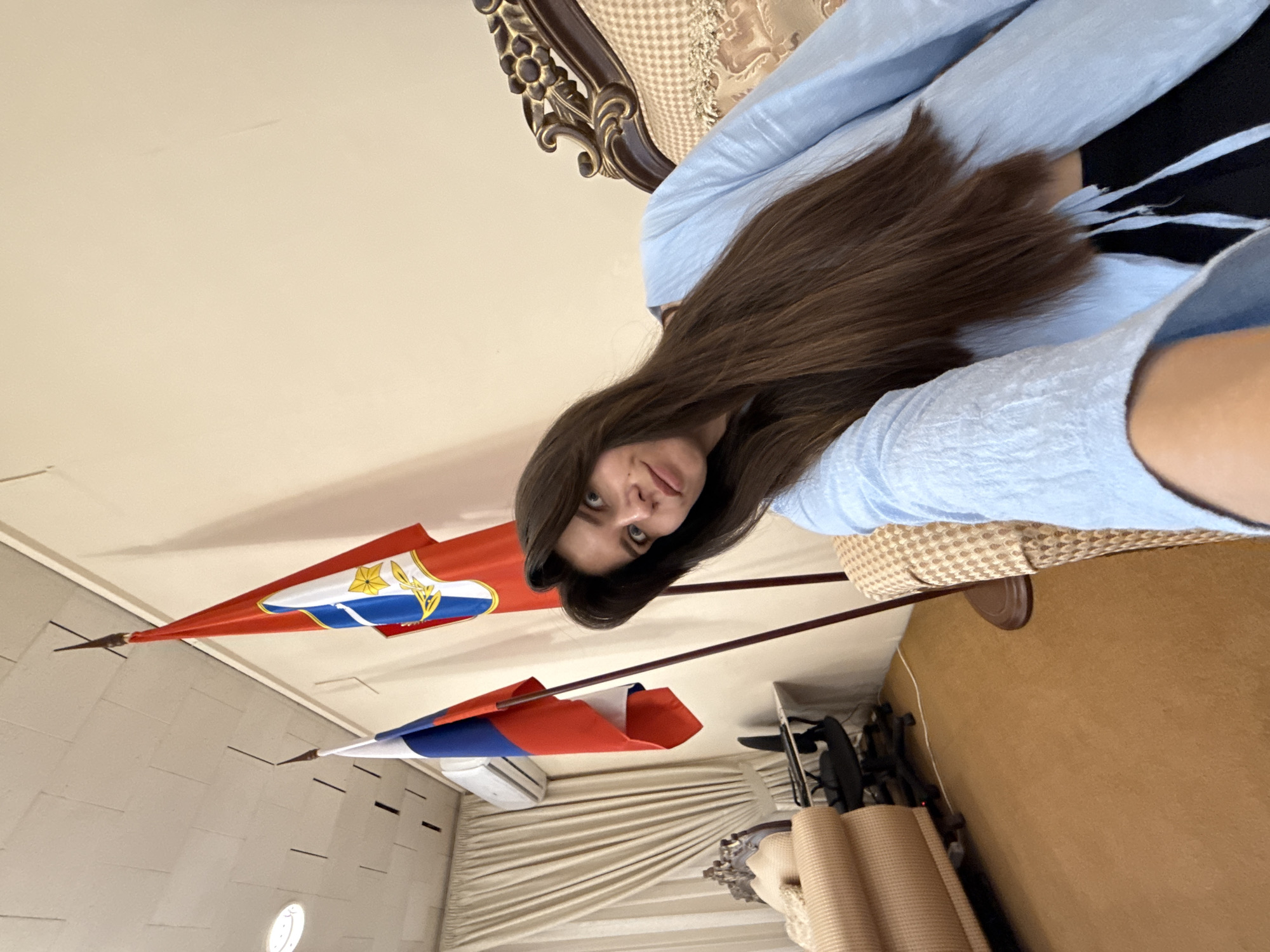 A young woman with long brown hair takes a selfie in a room with two flags, one red, white, and blue, and the other red with a coat of arms. She is wearing a light blue oversized shirt and black pants, and is seated on a couch with ornate wooden trim. Behind her are beige curtains, an air conditioner, and a desk with a chair. The floor is carpeted in a tan color.