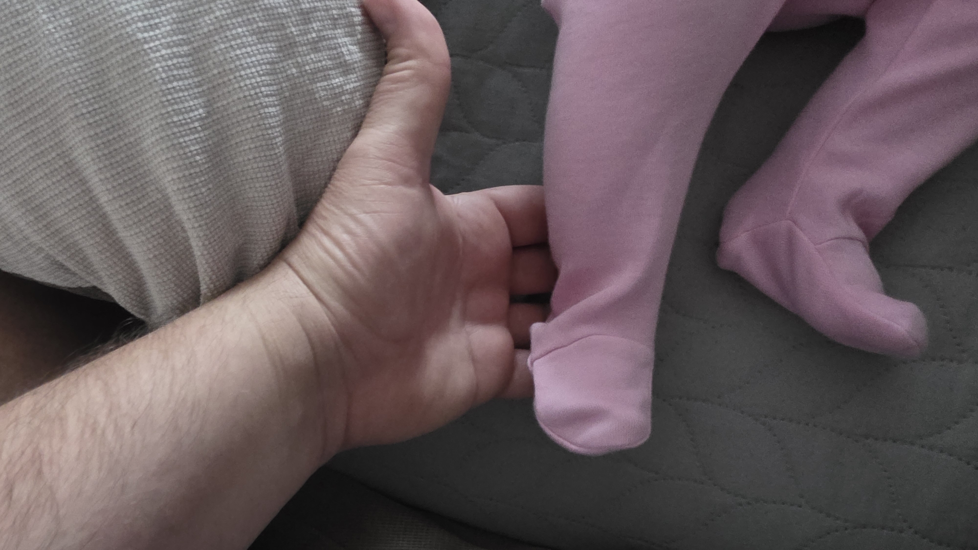 A close-up shot shows a man's hand gently holding the left leg of a baby wearing pink footed pajamas. The baby's right leg is also visible, positioned slightly behind the left leg. The hand is also holding a beige-colored cushion. The setting appears to be indoors, likely on a sofa or bed, judging by the grey quilted fabric in the background. There is no information available in the image to determine the location as Blagoevgrad, Bulgaria, or any other geographical location.