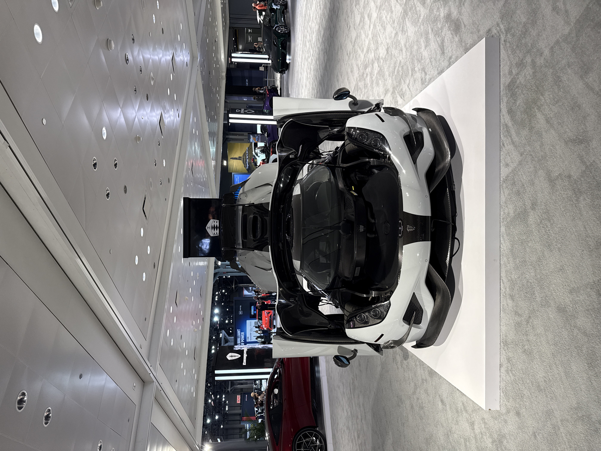 The image depicts a white and black Koenigsegg supercar on display, likely at an auto show or exhibition. The car is elevated on a white platform in a large indoor space, with the front hood open, revealing its engine and mechanical components. The location is inside a convention center, identifiable by the patterned ceiling with recessed lighting. The background shows other displays and vehicles, suggesting a car show or auto exhibition. People are visible in the background, indicating that the exhibition is open to the public. The overall environment is well-lit and modern, typical of a convention center. It is difficult to pinpoint the exact location in New York City without additional landmarks or signage.