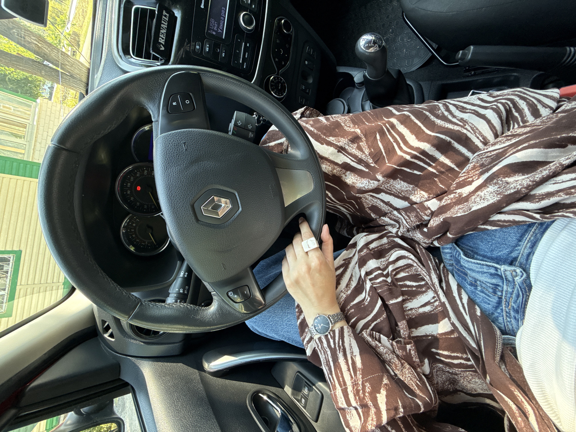 The image shows the interior of a car, focusing on the driver's side. The steering wheel, dashboard, and gear stick are visible. A person's hands are on the steering wheel, and they are wearing a watch and a ring. The person is wearing a patterned shirt and blue jeans. The car's interior appears to be clean and well-maintained. Outside the car, a building with green trim and trees are visible, suggesting an outdoor setting. The location is identified as Aksay, Kazakhstan, implying the car is parked or driving in this city.