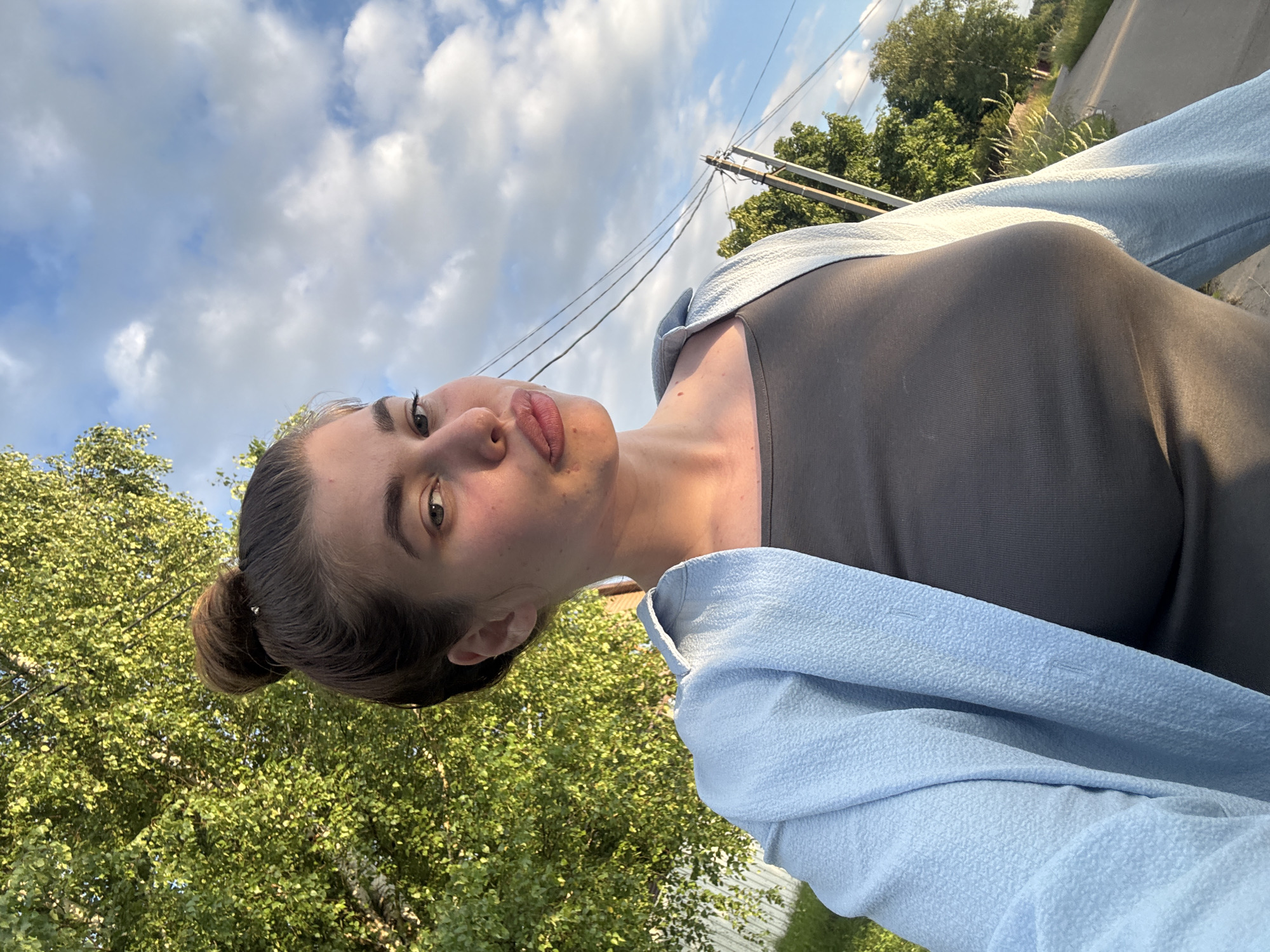 This is a portrait of a young woman with her hair in a bun, taken outdoors. She is looking towards the camera with a slight pout. She is wearing a grey top and a light blue, textured cardigan. The background consists of lush green trees, a bright blue sky with scattered white clouds, and utility poles with wires. The lighting suggests it is late afternoon or early evening, with the sun casting a warm glow on her face. The setting appears to be a rural or suburban area, possibly in Мещёры, Russia, given the natural landscape and power lines. The overall mood seems relaxed and casual. There is no visible text in the image.