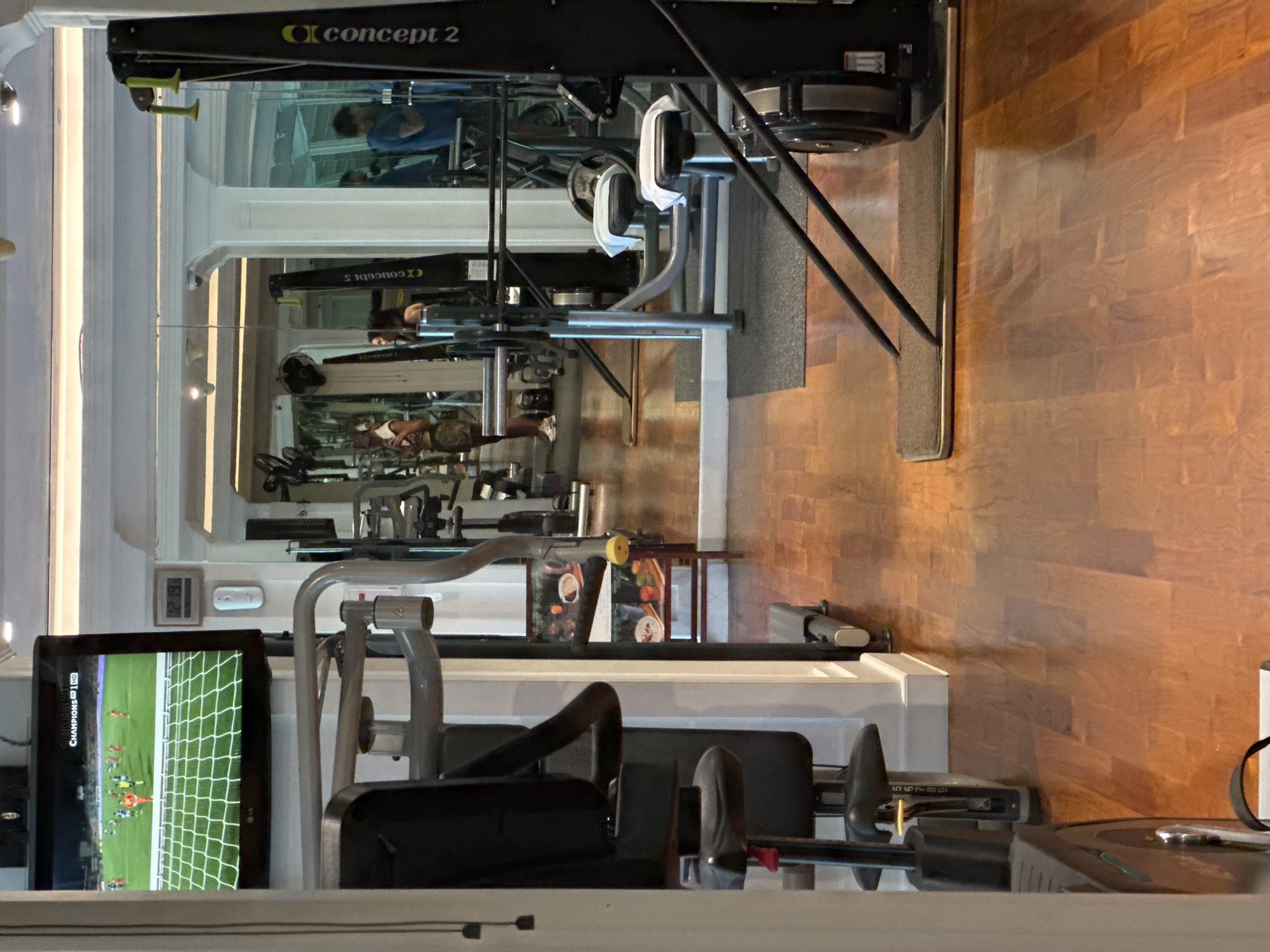 The image depicts a gym in Kuta Selatan, Indonesia. A Concept2 rowing machine is visible in the upper part of the frame. The gym has a mirror running along one wall. Several weight machines are present, and a television is visible in the lower left. The floor is made of wood. People are present in the mirror's reflection.