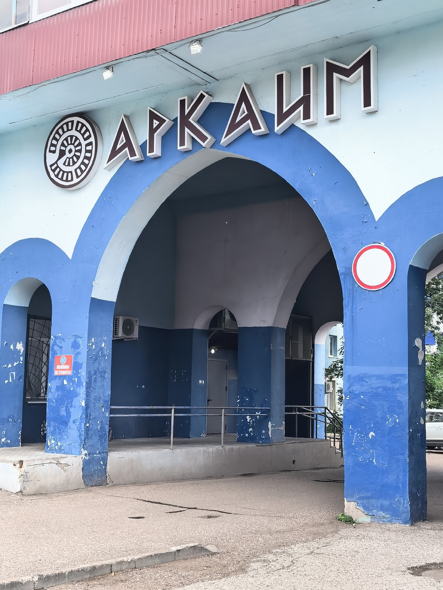The image shows an outdoor view of a building with blue arches and a red roof. On the building's facade, large, brown and white three-dimensional letters spell out "АРКАИМ" (Arkaim). To the left of the letters is a circular emblem with a spiral design. A red and white circular sign with a horizontal red bar across a white circle is affixed to one of the blue arch pillars. Below the arches, a paved area is visible, with a concrete curb in the foreground. The building appears to be a public or commercial establishment. The presence of the name "Arkaim" suggests a connection to the archaeological site and museum of the same name, which is located in the Chelyabinsk Oblast, Russia, not Sterlitamak. However, without further information, it is impossible to definitively state the specific location.