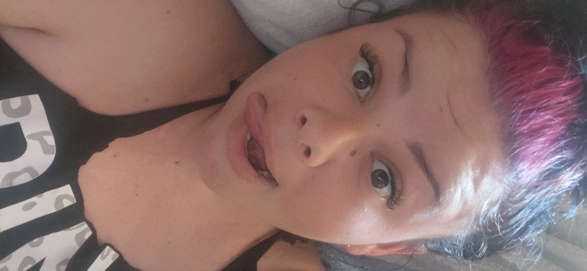 A woman with pink-streaked black hair and dark eyes is lying down, looking up at the camera. She is wearing a black t-shirt with a white and grey leopard-print pattern that spells out "VIVID". Her tongue is sticking out of her mouth. The image appears to be taken indoors, possibly in a bedroom, with soft lighting. The woman's expression is playful. There are no discernible environmental cues to indicate the time of day or weather. The setting is likely a domestic space in Marynivka, Ukraine.