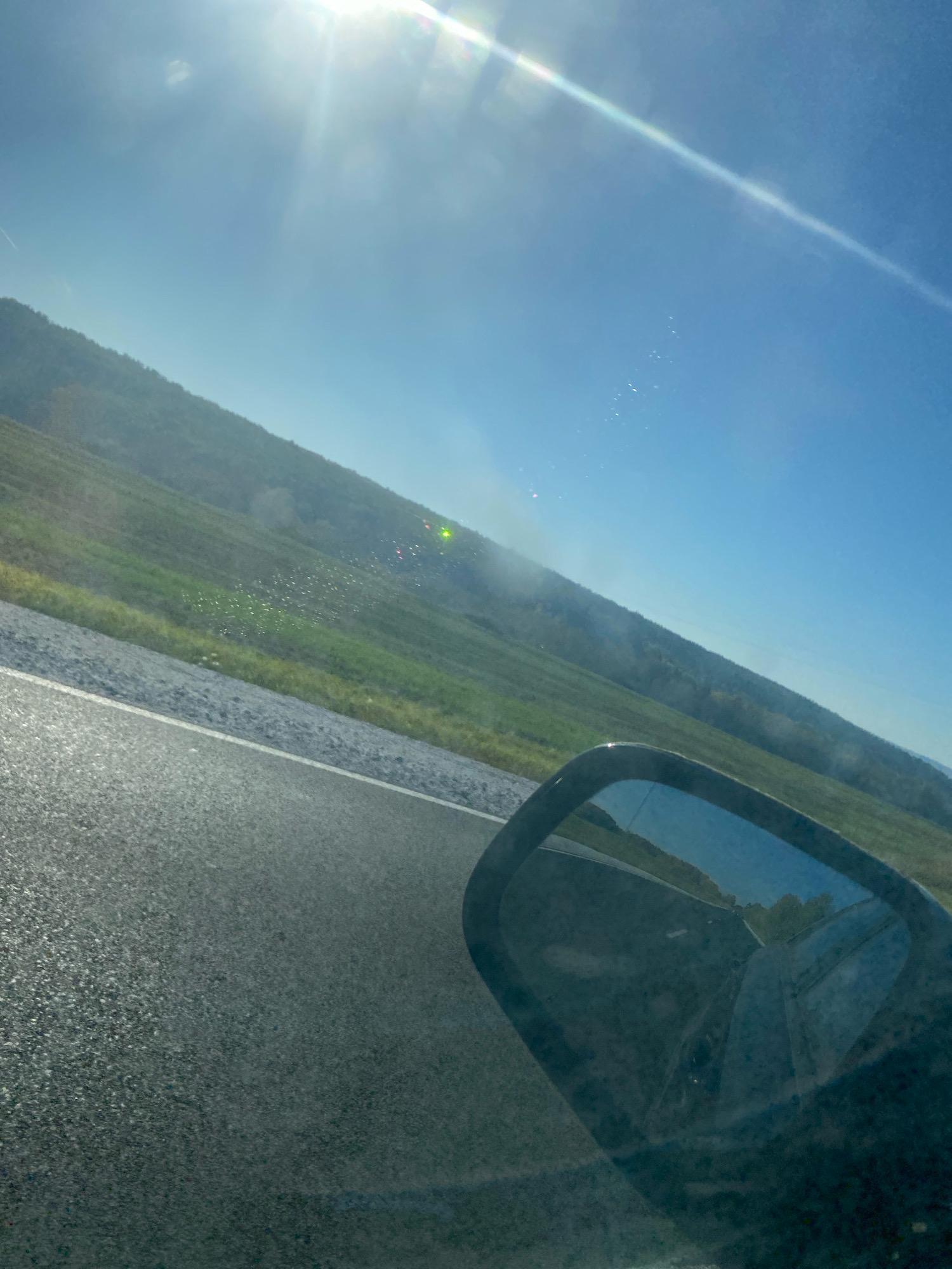 The image, taken from a vehicle on a road, shows a sunlit scene with a bright sun in the upper left corner casting lens flare. A green, grassy hillside slopes upwards from the right side of the frame towards the left. The road in the foreground is asphalt, with a grey gravel shoulder. In the distance, there are some small, scattered red and green lights on the hillside, possibly from vehicles or lights on the terrain. A side-view mirror is visible on the right side of the frame, reflecting the road and the sky. There is a hint of smoke or mist rising from the hillside in the middle ground. The sky is a clear blue. This image does not contain any recognizable landmarks or features that would allow for its identification to the city of Pacha, Russia.