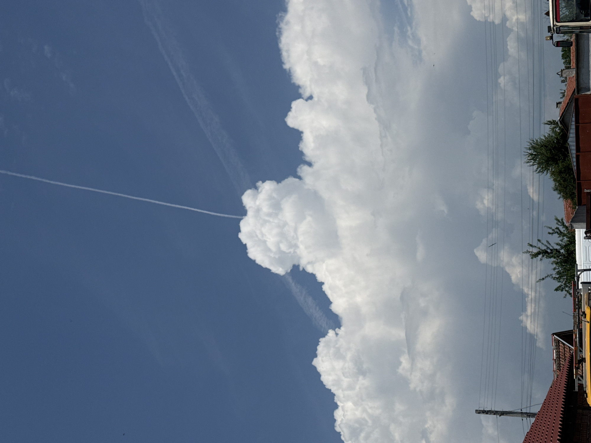This image depicts a daytime outdoor scene in Sarnevo, Bulgaria, characterized by a vibrant blue sky filled with white cumulus clouds. A prominent band of dense, bright white cumulus clouds stretches across the middle of the frame, with one large, central formation rising significantly higher into the blue sky. A distinct, thin white contrail extends diagonally from the upper left corner, passing behind the apex of this central cloud formation. A second, fainter contrail is horizontally visible in the upper right quadrant. Scattered small, dark specks, indicative of birds or insects, are present against the blue sky. The lower section of the image shows elements of a built environment: several terracotta-tiled rooftops are partially visible across the bottom, alongside the top portion of a yellow vehicle and a section of a white vehicle. A wooden utility pole on the left supports multiple dark power lines that traverse the lower middle of the frame horizontally. Green foliage, including trees and shrubs, is visible above the rooftops in the lower half of the image.