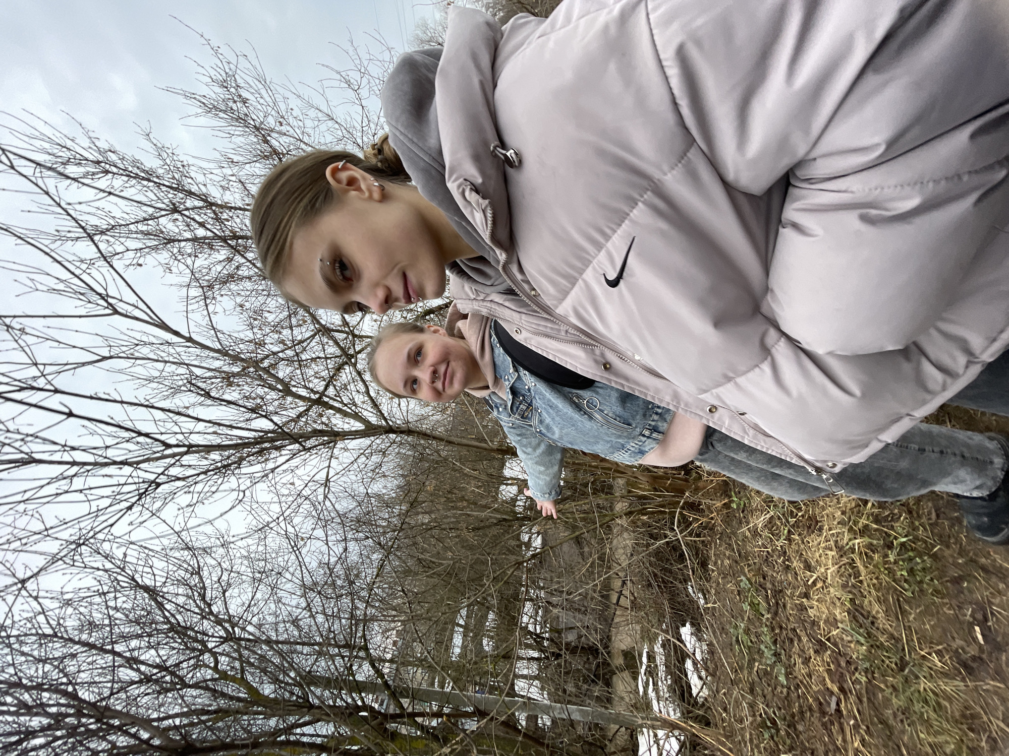 Two women stand outdoors in a landscape dominated by bare trees, suggesting a cold season, possibly late autumn or early spring. The location is Фоки, Russia. The woman in the foreground wears a light pink puffer jacket with a gray hood and a visible Nike logo. She has multiple piercings in her left ear and an eyebrow piercing on the same side. The second woman behind her wears a denim jacket over a lighter-colored hoodie. Both are partially visible, suggesting they're standing close together. The ground has patches of snow and dead grass, hinting at recent snowfall or melting snow. A metal railing is visible in the background.