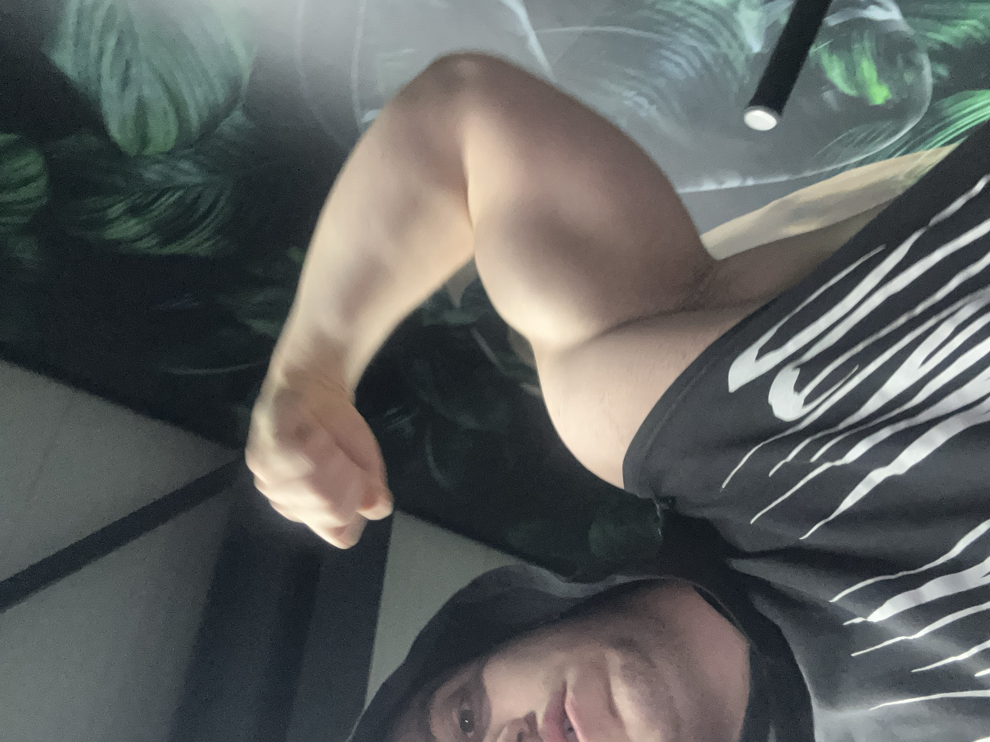 A selfie image captures a man wearing a dark hooded sweatshirt and a black tank top with white, zebra-like stripes. His left arm is flexed, showcasing his bicep. The background features a screen displaying a wallpaper of green leaves. The photograph provides no information that can place this image in Burgas, Bulgaria.