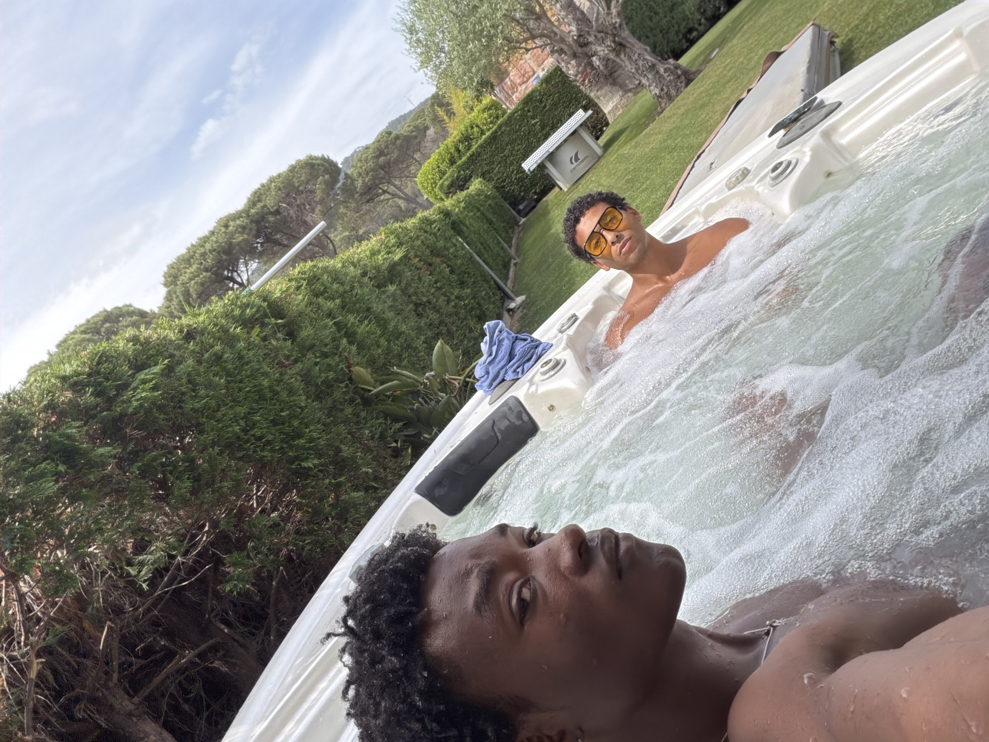 Two young Black men are in a white hot tub. The hot tub is in a yard with green grass and hedges. One man is wearing yellow sunglasses. A blue towel is on the edge of the hot tub. A white ping pong table is behind the hot tub, near a tree. The location is Sant Andreu de Llavaneres, Spain.