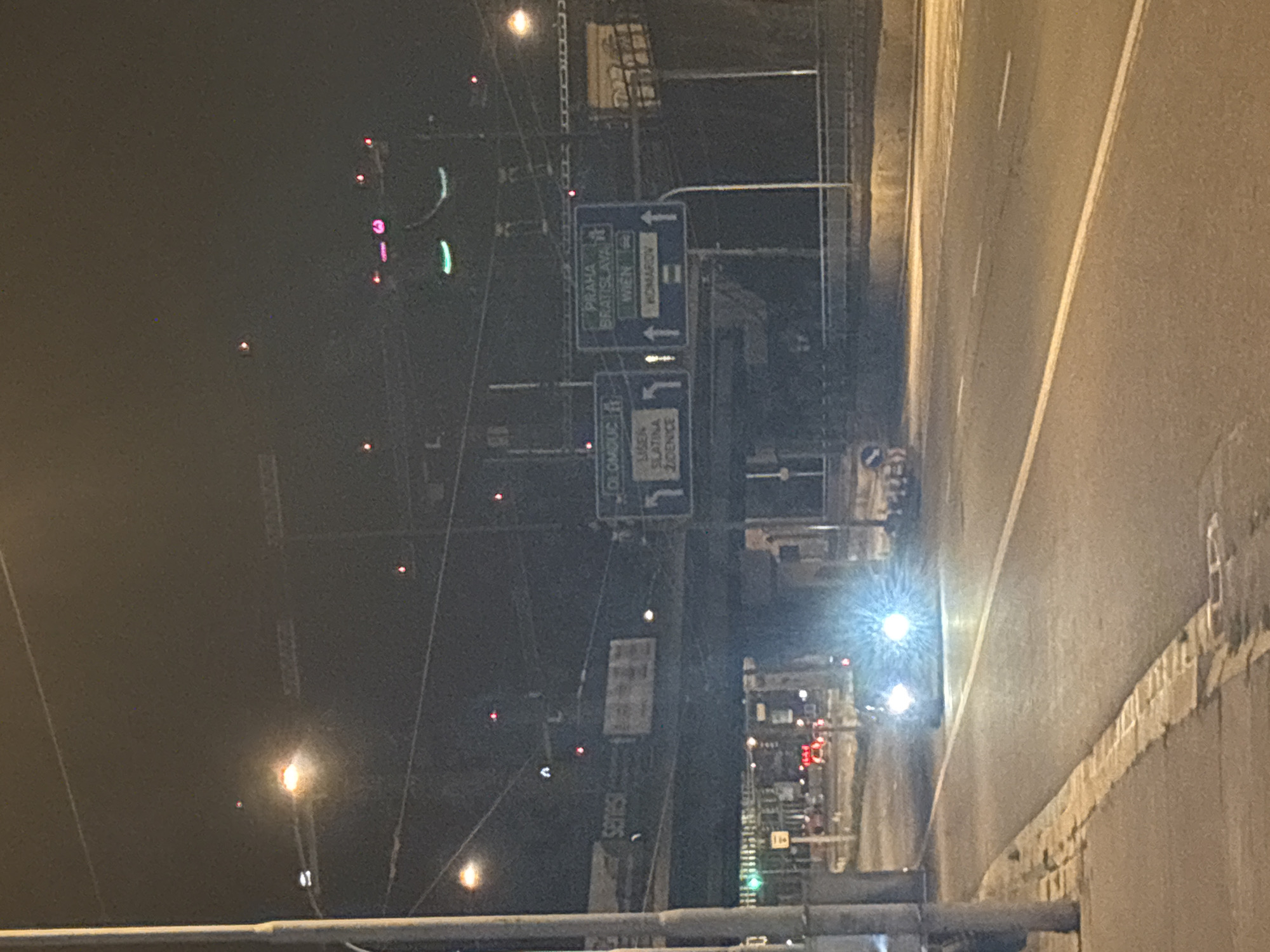 The image captures a night scene on a road in an urban environment. A car with its headlights on is the primary subject, positioned in the center of the frame. The car's headlights are very bright, creating a lens flare effect. The road is paved and has white lane markings. To the left of the road, there is a sidewalk with a curb and a pole. Above the road, an elevated structure, possibly a bridge or overpass, is visible. On this structure, there are road signs indicating directions to cities such as OLOMOUC, PRAHA, BRATISLAVA, WIEN, and KOMAROV. Some lights are also visible on the elevated structure and in the background. The overall atmosphere is dark, illuminated by artificial lights.