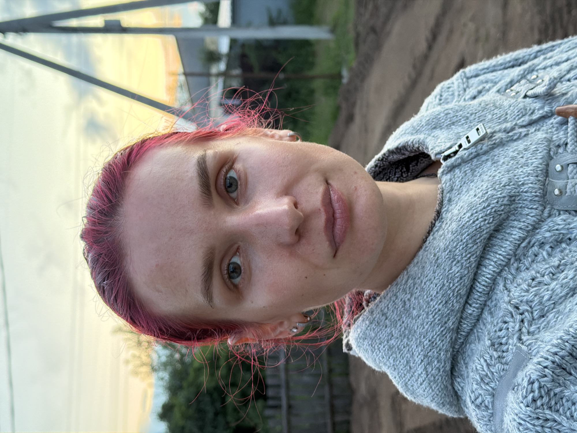 A woman with bright pink hair and wearing a light grey hooded sweater is pictured in a close-up selfie taken outdoors in Chaikovsky, Russia. The background shows a dirt path, wooden fencing, trees, and a utility pole against a twilight sky. The woman's face is the primary focus, with her eyes looking directly at the camera.