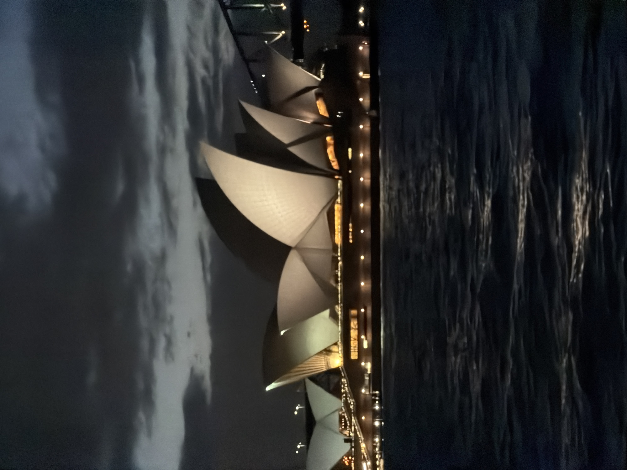 The image depicts the Sydney Opera House at night, illuminated against a dark, cloudy sky. The iconic white sails of the opera house are lit, with warm yellow light emanating from within the structure and along its base. The water in the foreground reflects the lights of the city and the moon. The Sydney Harbour Bridge is visible in the background, also lit up with its own lights. The overall scene is one of a majestic landmark bathed in artificial light under a dramatic night sky.