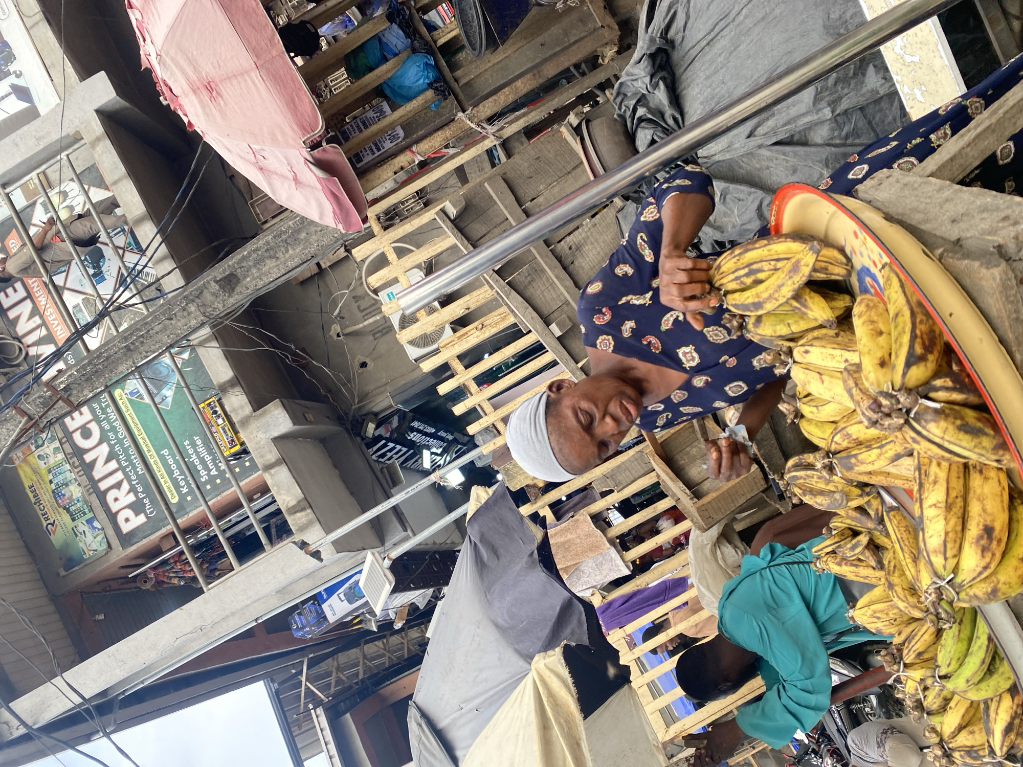 An eye-level, tilted shot depicts an outdoor marketplace in Lagos, Nigeria. An African woman wearing a dark blue, patterned top and a white head covering is seen examining a bunch of plantains. The plantains are in an oval, yellow tray with a blue and red rim. A man in a teal shirt stands next to the woman, also examining the plantains. A motorcycle is visible under the plantains and the wooden crate in which they are being sold.
The background shows a jumble of wooden pallets, tarps, and other market stalls. In the distance, a pink umbrella is tilted left, and a sign for "PRINCE VINE" is visible. Power lines crisscross the sky.