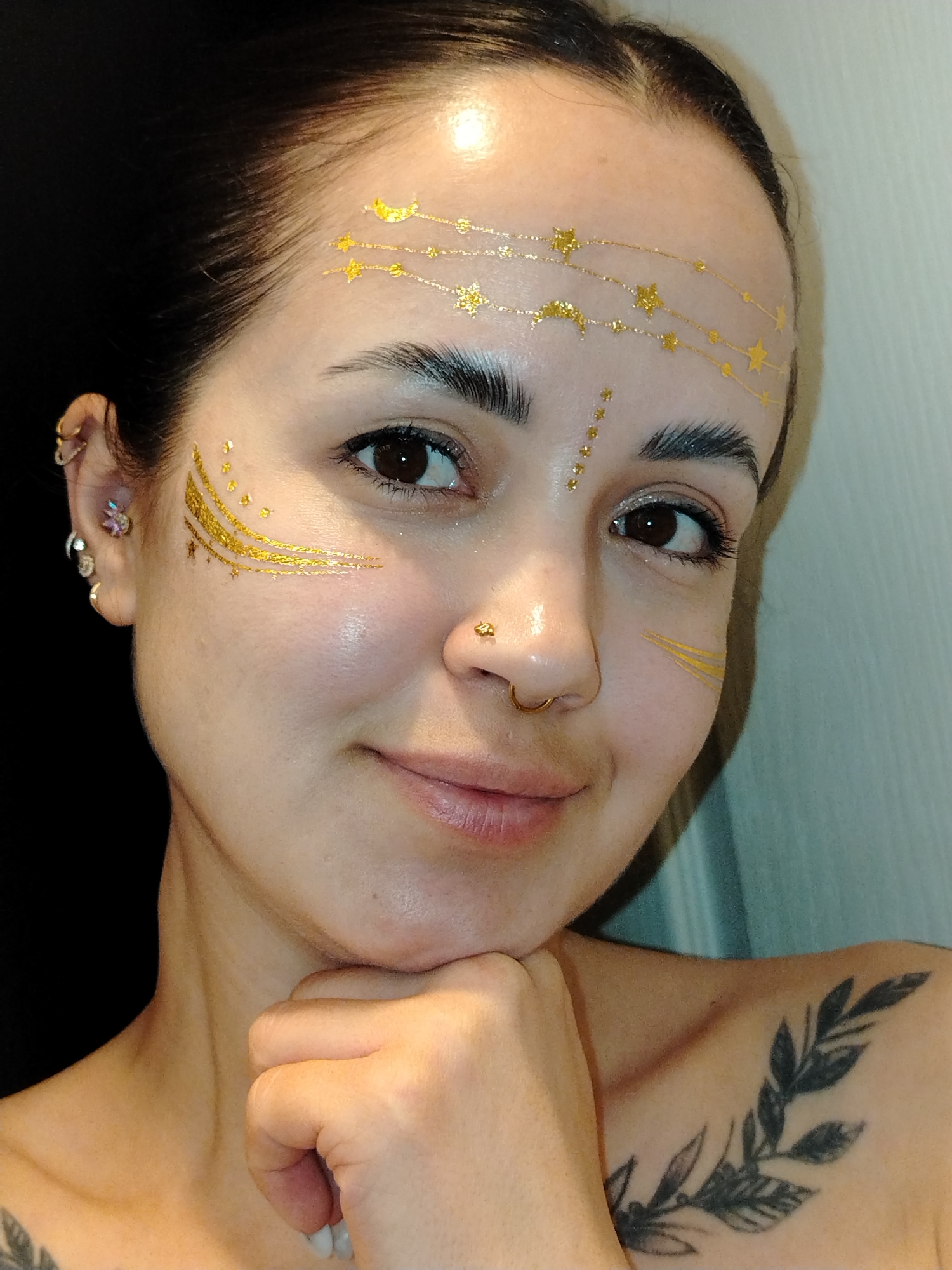 A close-up portrait captures a woman in an indoor setting, likely in Noviy, Russia, showcasing intricate gold facial decorations. She appears to be in her 20s or 30s, with dark hair neatly pulled back from her face. Her forehead is adorned with delicate gold temporary tattoos depicting constellations, including stars and crescent moons, connected by dotted lines. Similar golden designs, resembling brushstrokes and small stars, embellish the areas around the outer corners of her eyes and upper cheekbones.
The woman has several facial piercings: a gold hoop through her septum, a small gold stud on the right side of her nose, and multiple earrings in her left ear, including a hoop, a stud, and a small, star-like cluster. Her dark eyebrows are well-groomed, and her brown eyes look directly at the camera with a subtle, friendly gaze. She has a hint of glittery eyeshadow on her eyelids.
She is depicted with a gentle smile, her chin resting on her lightly fisted right hand. A dark-ink tattoo of a leafy branch design is visible on her right shoulder/collarbone area. The background is mostly dark on the left, possibly a dark wall or object, contrasting with a lighter, subtly textured vertical surface on the right. The lighting is bright and direct, highlighting the metallic sheen of the face art and her glowing skin. The image conveys a sense of artistic expression or readiness for a celebratory occasion. No visible text is present in the image.