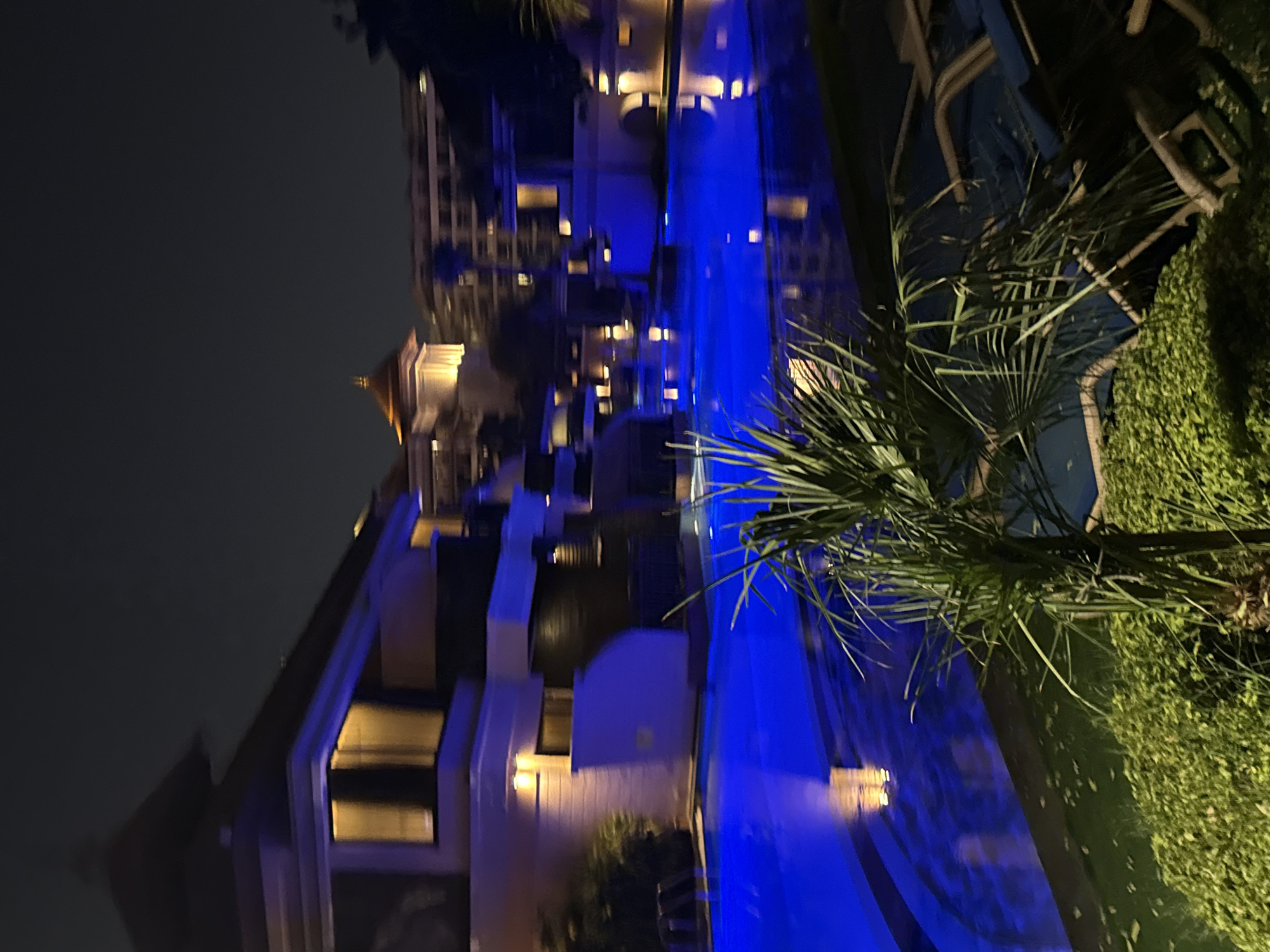 A nighttime scene shows a resort with a large, brightly lit blue swimming pool. Buildings with illuminated windows are visible behind the pool, as well as lush green foliage and palm trees in the foreground. The architecture suggests a luxurious resort, possibly in a warm climate, consistent with Dubai, United Arab Emirates, known for its modern hotels and leisure facilities. There are no people or animals visible in the image. The overall impression is one of a serene and inviting evening ambiance at a high-end establishment.