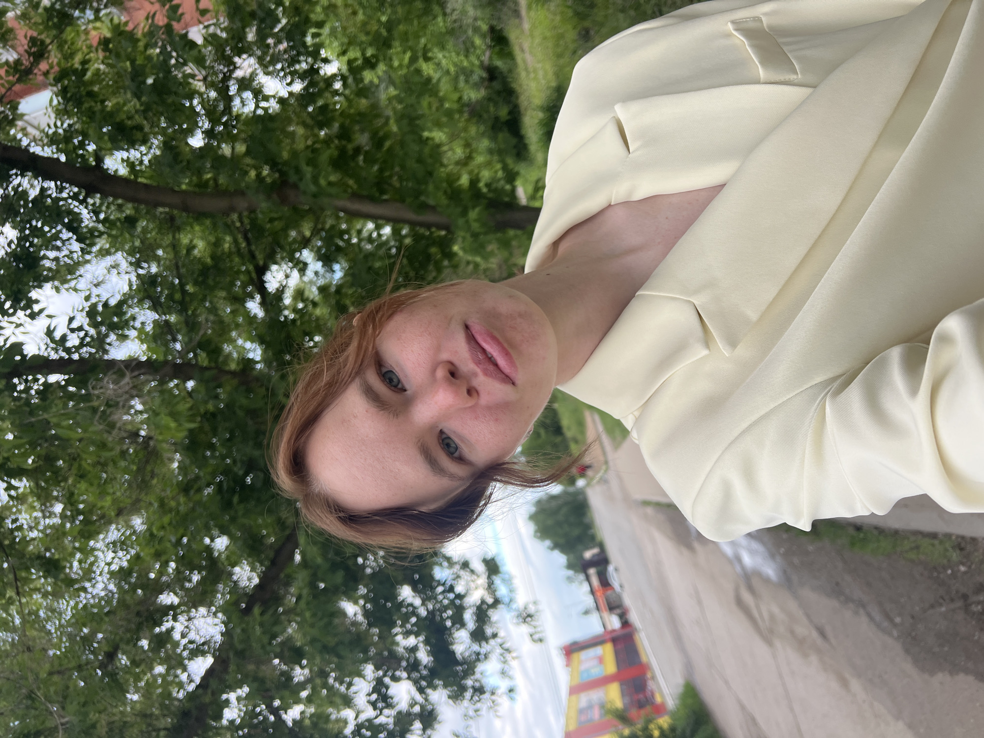 The image presents a close-up selfie of a young individual, likely female, with reddish-brown hair and a fair complexion exhibiting visible freckles across the nose and cheeks. Her eyes are light-colored (appearing blue or green), and she maintains a neutral expression while looking directly at the camera. She is dressed in a light cream or pale yellow blazer, discernible by its lapels and visible stitching on the right side of the frame.
The background places the scene outdoors on a street in what is described as Chaikovsky, Russia. To the right and behind the individual, a significant portion of the frame is filled with lush green foliage from trees, including a distinct tree trunk, suggesting a park or a heavily treed roadside area. To the left, a paved road or street is visible, with dark, damp patches, indicating a wet surface. Across this road, a prominent commercial building with distinct red and yellow paneling is clearly visible. Further down the street, additional structures and a white vehicle are discernible. The sky above is predominantly overcast with soft cloud formations. The environment combines elements of urban infrastructure with natural green spaces.