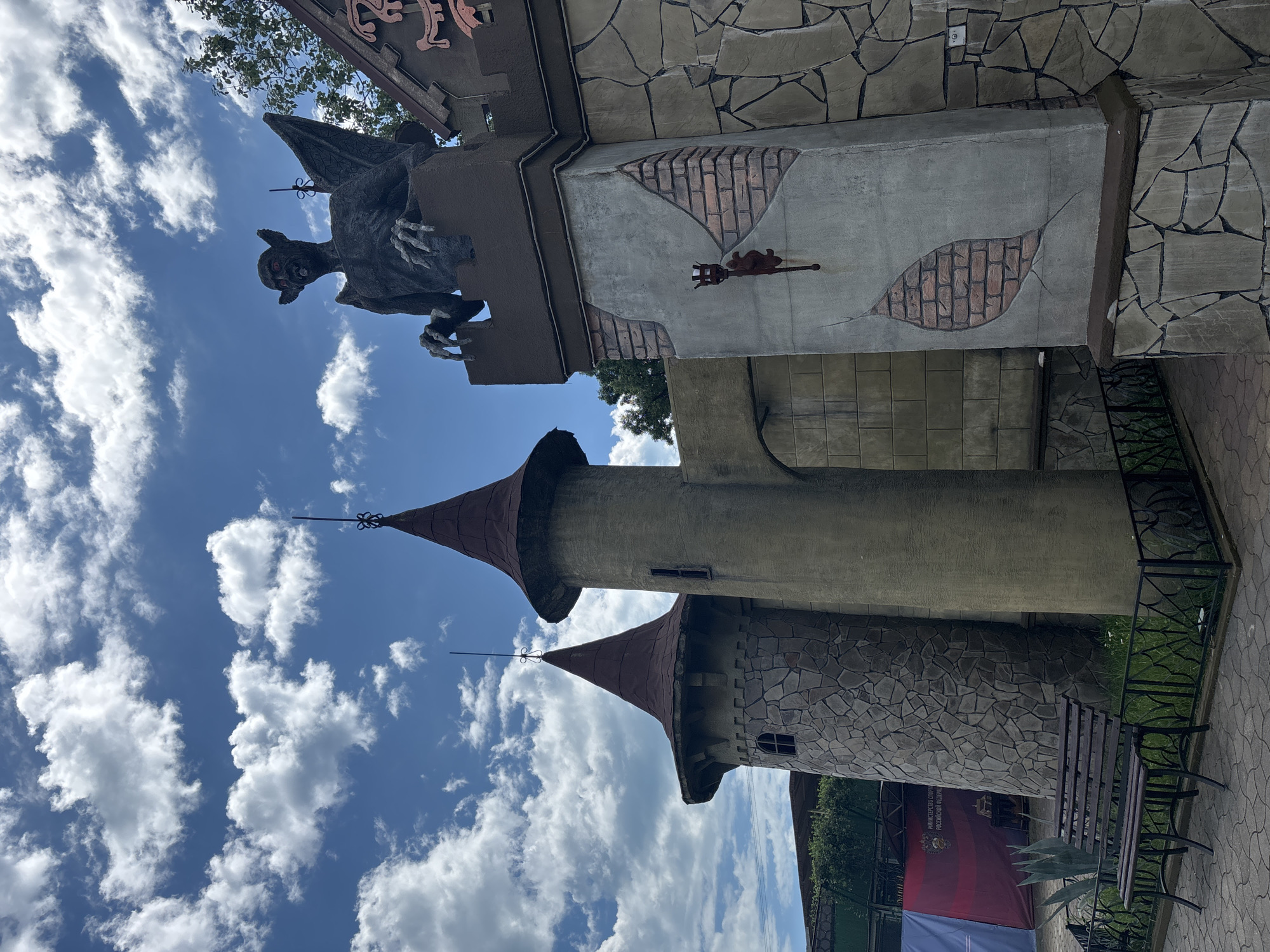 A stone-like building, resembling a castle with conical roofs, is seen under a bright blue sky with scattered white clouds. A dark statue of a winged creature with red eyes and sharp claws sits atop a battlemented section of the building on the right. A rustic torch fixture is mounted on the wall below the statue. To the left, two towers of the castle-like structure are visible, one made of rough stone blocks and the other a smooth, light brown. A wooden bench sits in front of a low black fence, with a paved walkway behind it. In the background, banners and trees are visible, suggesting an outdoor park or recreational area. The scene appears to be in Sochi, Russia, as indicated by banners with Russian text.