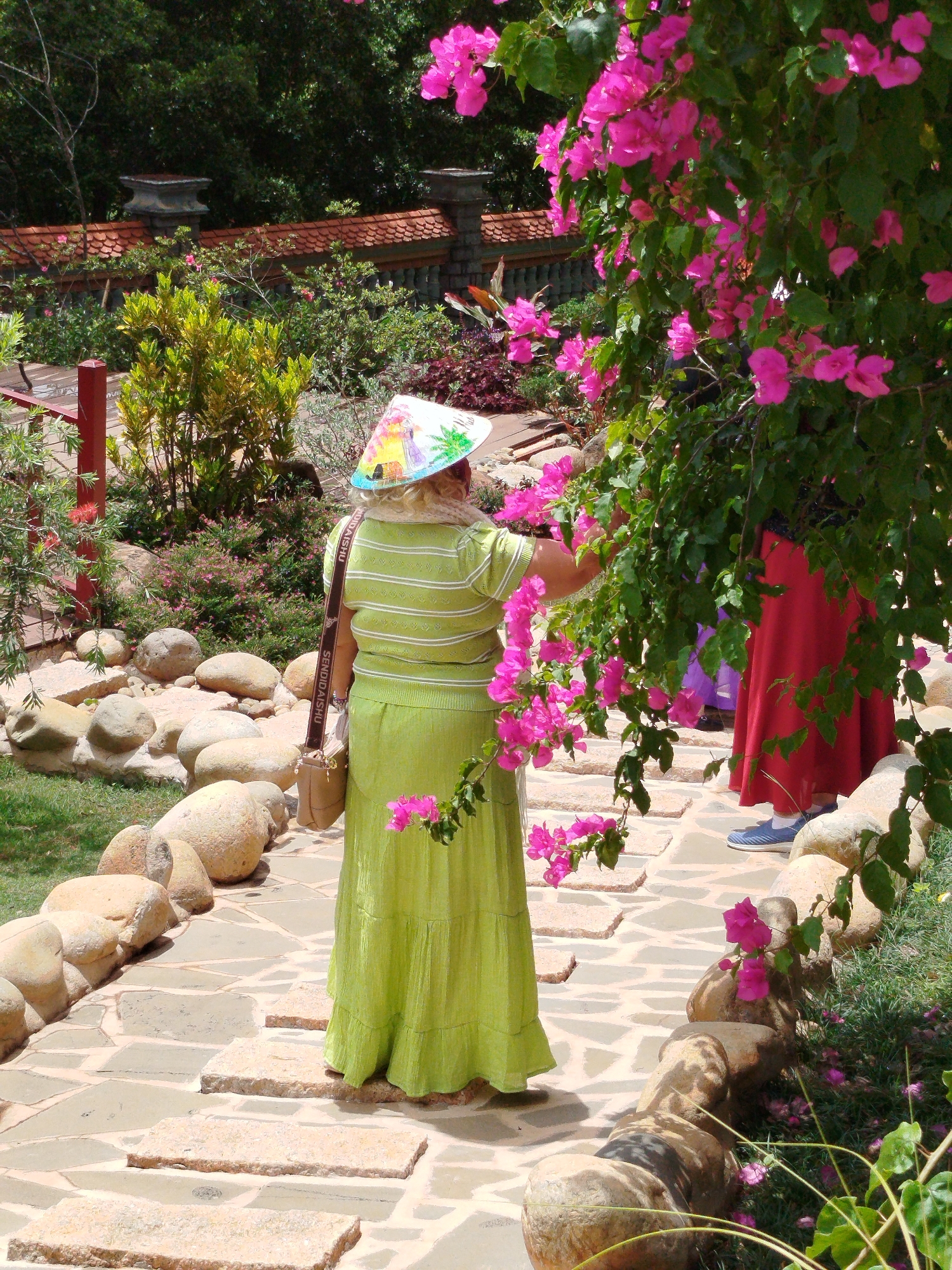 A medium outdoor shot shows a woman in a green outfit and straw hat standing on a stone walkway and examining a pink flowering bush.
The woman is facing away from the camera, wearing a green skirt and shirt, and a straw hat with a landscape painting. She has a purse on her shoulder and is reaching out to touch a pink flower.
Behind her, another person in a red dress and blue shoes is also looking at the flowers. The walkway is made of irregular stones and is lined with large, round rocks. The background consists of trees, bushes, and a low brick wall.
The location is the grounds of the Truc Lam temple in Da Lat, Vietnam.