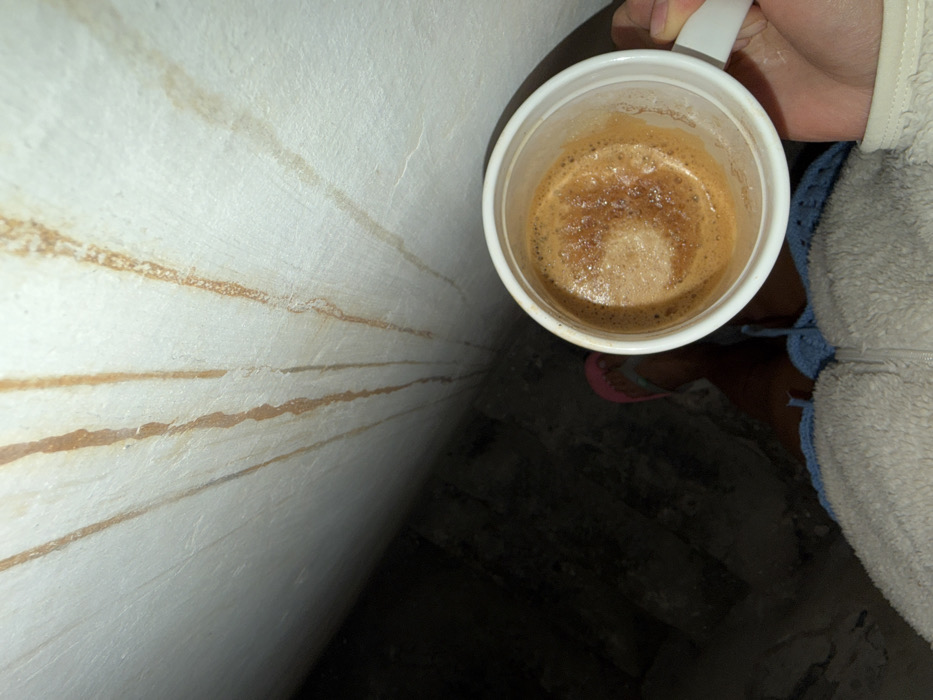 The image depicts a first-person view of a person holding a white mug containing a dark brown beverage with froth, likely coffee. The person's right hand grips the mug handle. The individual wears light-colored, textured outerwear, possibly fleece, with a visible zipper, blue woven shorts, and pink flip-flops. Dominating the upper portion of the frame is an off-white wall exhibiting multiple vertical reddish-brown streaks, consistent with rust or water damage. The lower left of the frame is a dark, indistinct area, possibly a shadowed floor or ground surface. No visual identifiers are present to determine the location within Tel-Aviv, Israel.