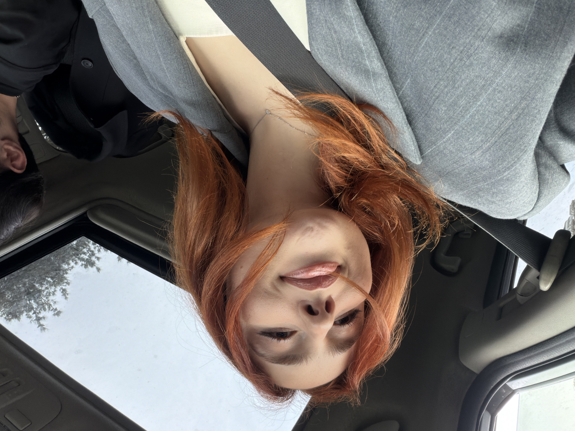 A woman with auburn hair is sitting in a car. She is wearing a gray jacket over a white top and a seatbelt. The car has a sunroof through which a view of a cloudy sky and trees can be seen. The location is likely Tbilisi, Georgia. There is another person, possibly a male, in the vehicle with her.