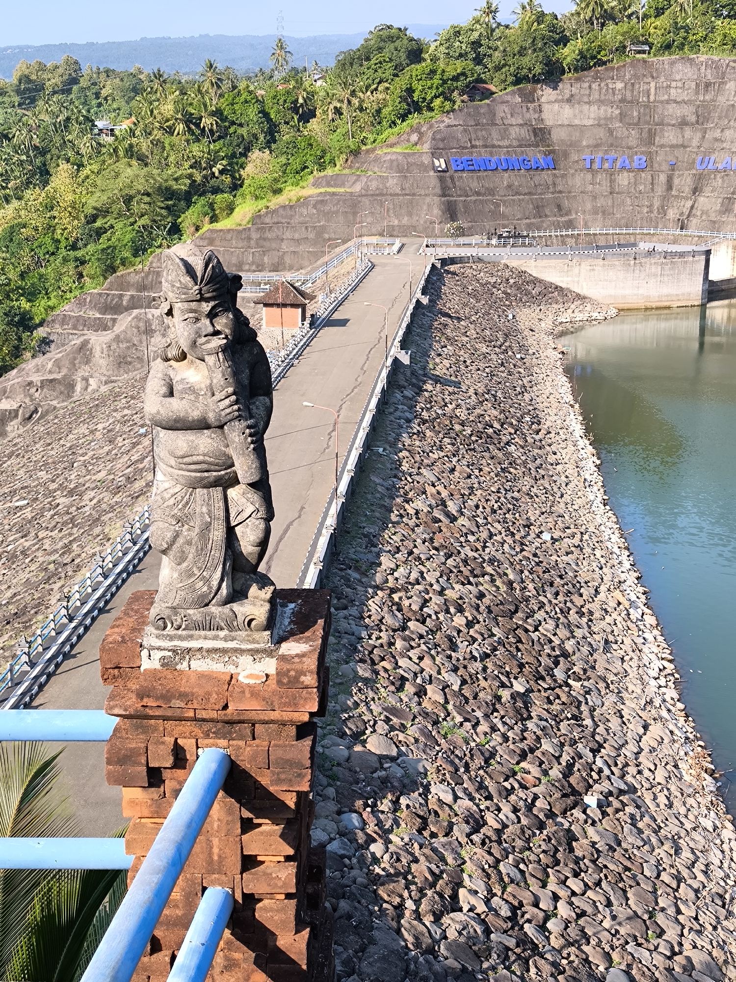 The image captures a detailed view of the Bendungan Titab-Ularan (Titab-Ularan Dam) located near Seririt, Buleleng Regency, Bali, Indonesia. In the immediate foreground, a weathered stone statue of a humanoid figure, sculpted in a traditional Balinese style, stands on a reddish-brown brick pedestal. The figure appears stout, with prominent facial features and stylized hair or headwear, and is holding a long, pipe-like object vertically in its hands. Blue painted metal railings are visible to the left and below the statue, indicating a vantage point from a bridge or walkway.
The midground is dominated by the massive concrete structure of the dam. A paved road stretches across the top of the dam wall, bordered by white railings and streetlights. Prominently displayed on the grey concrete face of the dam, large blue three-dimensional letters spell out "BENDUNGAN TITAB - ULA". A body of green-tinged water, likely the reservoir, is visible to the right of the road, with a steep embankment of large, irregularly shaped rocks forming its edge. Beyond the dam, the terrain rises into lush green hills densely covered with tropical vegetation, including numerous palm trees. Distant mountains or hills are visible under a clear, bright blue sky. The scene is illuminated by strong sunlight, casting distinct shadows, indicative of daytime. No human figures are explicitly visible in the frame.