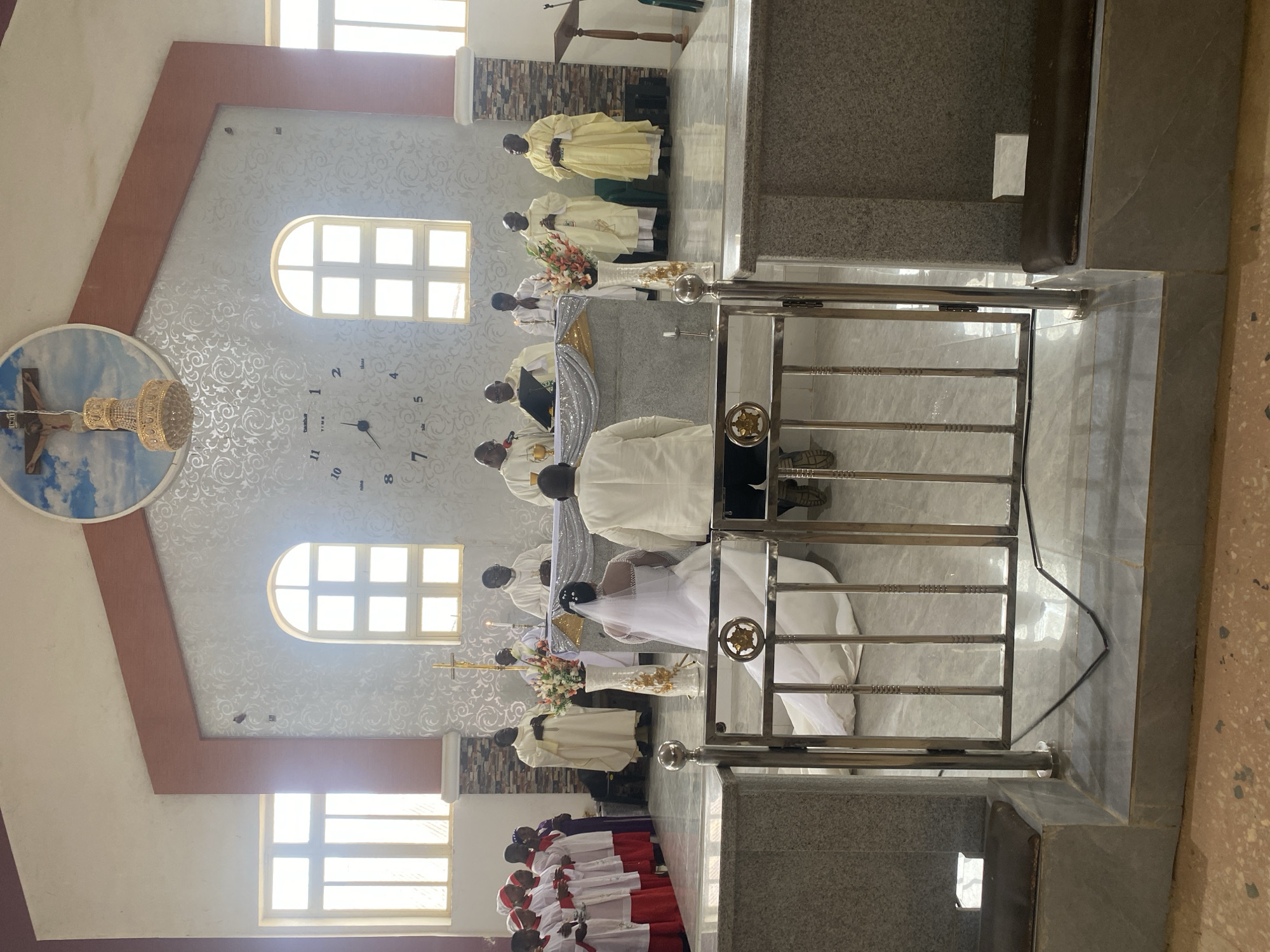 In a church, likely in Jos, Nigeria, a wedding is taking place. A bride in a white gown and veil kneels alongside a groom in white. Officiating clergy dressed in white and gold vestments face the couple. Behind the clergy are other figures, including clergy in yellow vestments and another group in red and white robes. The wall behind them has three windows and a round wall-hanging with a religious image. A decorative clock is also on the wall. A metal railing is in the foreground.