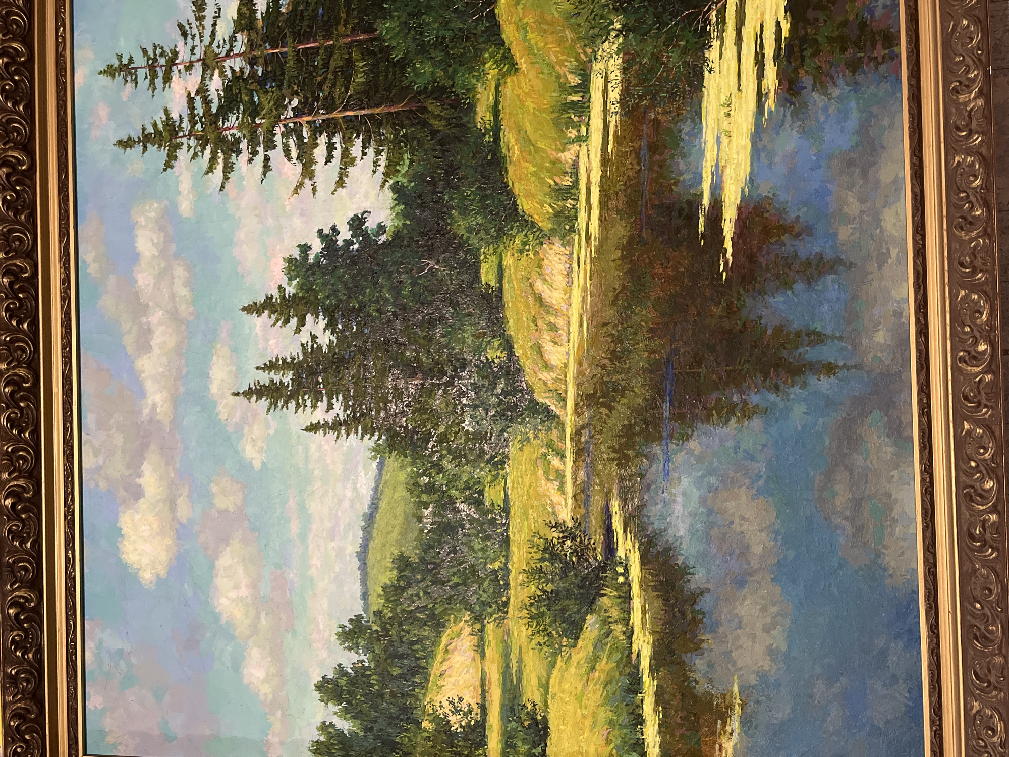 The media file displays a vibrant oil painting of a serene natural landscape, enclosed in an ornate gold frame. The painting is oriented horizontally.
The central subject is a tranquil body of water, likely a lake or a wide river, occupying the right half of the canvas. The water vividly reflects the clear, blue sky above, interspersed with soft, scattered white and light gray clouds. Along the left bank of the water, a dense forest of tall, dark green pine or fir trees stands prominently. Beyond the trees, rolling hills covered in lush green and sunlit yellow-green foliage are visible, extending into the background. Some trees, possibly willows or similar, have long, bright yellow-green branches hanging low over the water, creating striking reflections.
The painting evokes a peaceful and tranquil atmosphere, emphasizing the beauty of nature. The time of day appears to be early morning or late afternoon, as suggested by the warm, bright light illuminating the grassy hills and shimmering on the water's surface, particularly on the right side. The sky is partly cloudy, contributing to a sense of a pleasant day. The artistic style features visible brushstrokes, giving the painting an impressionistic quality with rich textures and vibrant colors. There are no human figures or animals depicted, nor any visible text within the painting. The location context provided is Новая Тура, Russia. The intricate, dark gold frame adds a classical touch to the artwork.