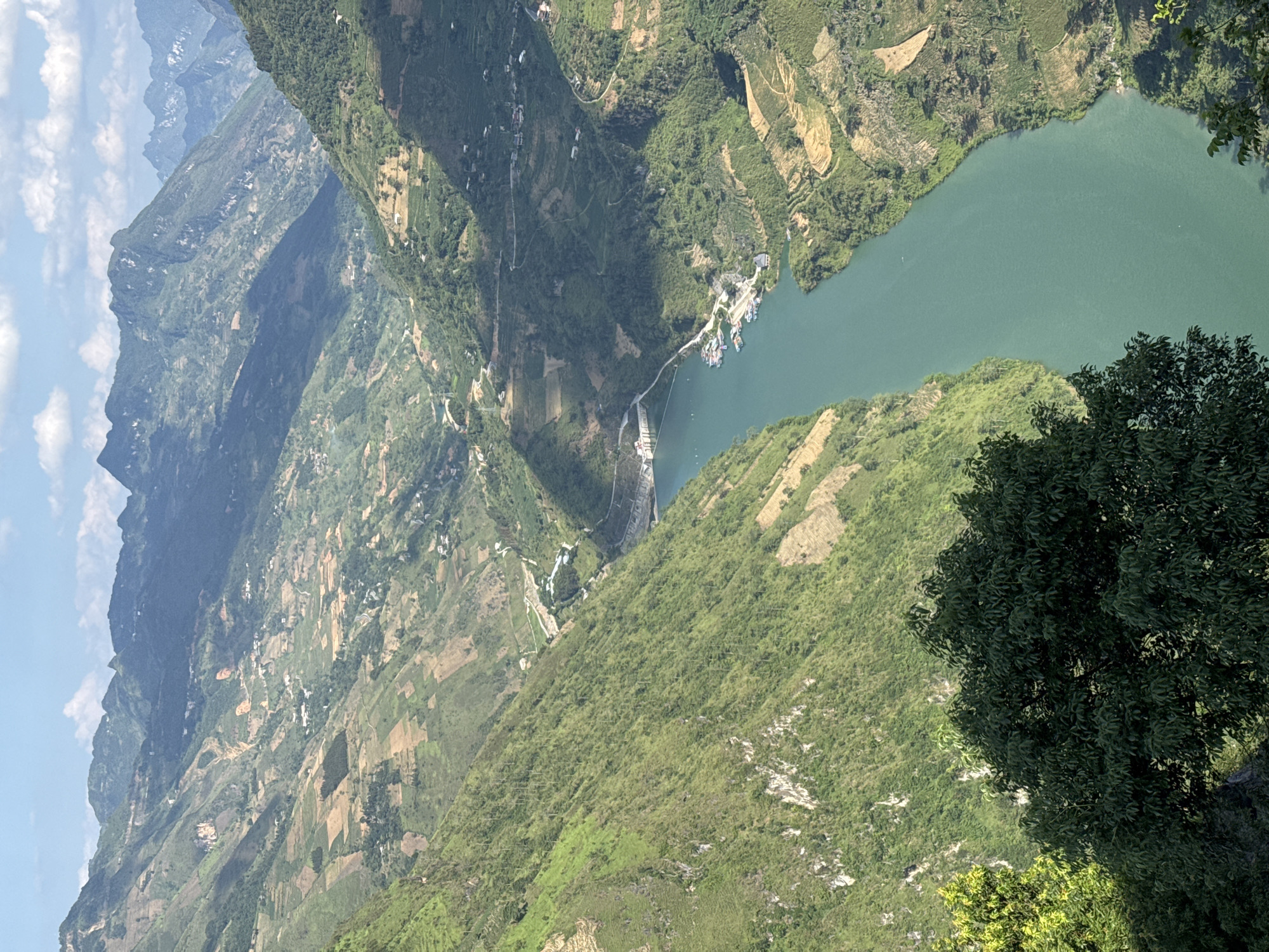 The image shows a steep, mountainous landscape in Đồng Văn Commune, Vietnam. A green river flows through a valley formed by the mountains. Terraced farming is visible on the slopes. A dam is present on the river, with docked boats nearby. The sky is blue with some clouds. Tree foliage is visible in the lower right corner of the image.