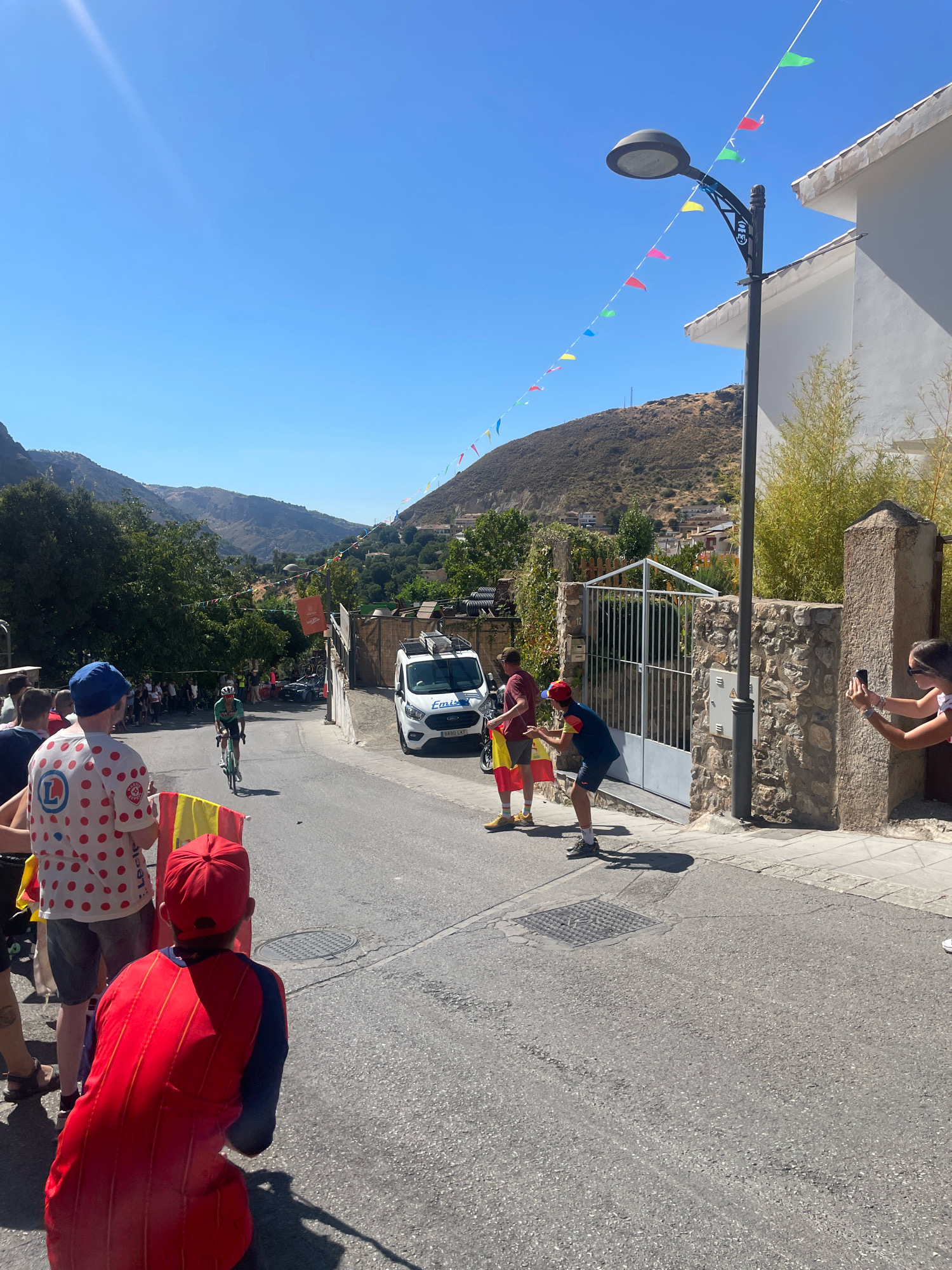 The image depicts a street scene in Güéjar Sierra, Spain, under a clear blue sky. A cyclist, wearing a green jersey and white helmet, is riding up a moderately sloped asphalt road. In the foreground, several spectators are gathered, some holding Spanish flags. One person in the front wears a white shirt with red polka dots and a blue hat. Another person in a red shirt and red cap is crouching. On the right side of the road, a white van with "Emisa" visible on its side is parked. A stone wall with a metal gate is also visible. Colorful triangular flags are strung across the street on wires, attached to a lamppost. In the background, the landscape features rolling hills covered with sparse vegetation and scattered buildings, characteristic of a mountainous region. The overall atmosphere suggests a public event, possibly a cycling race or festival.