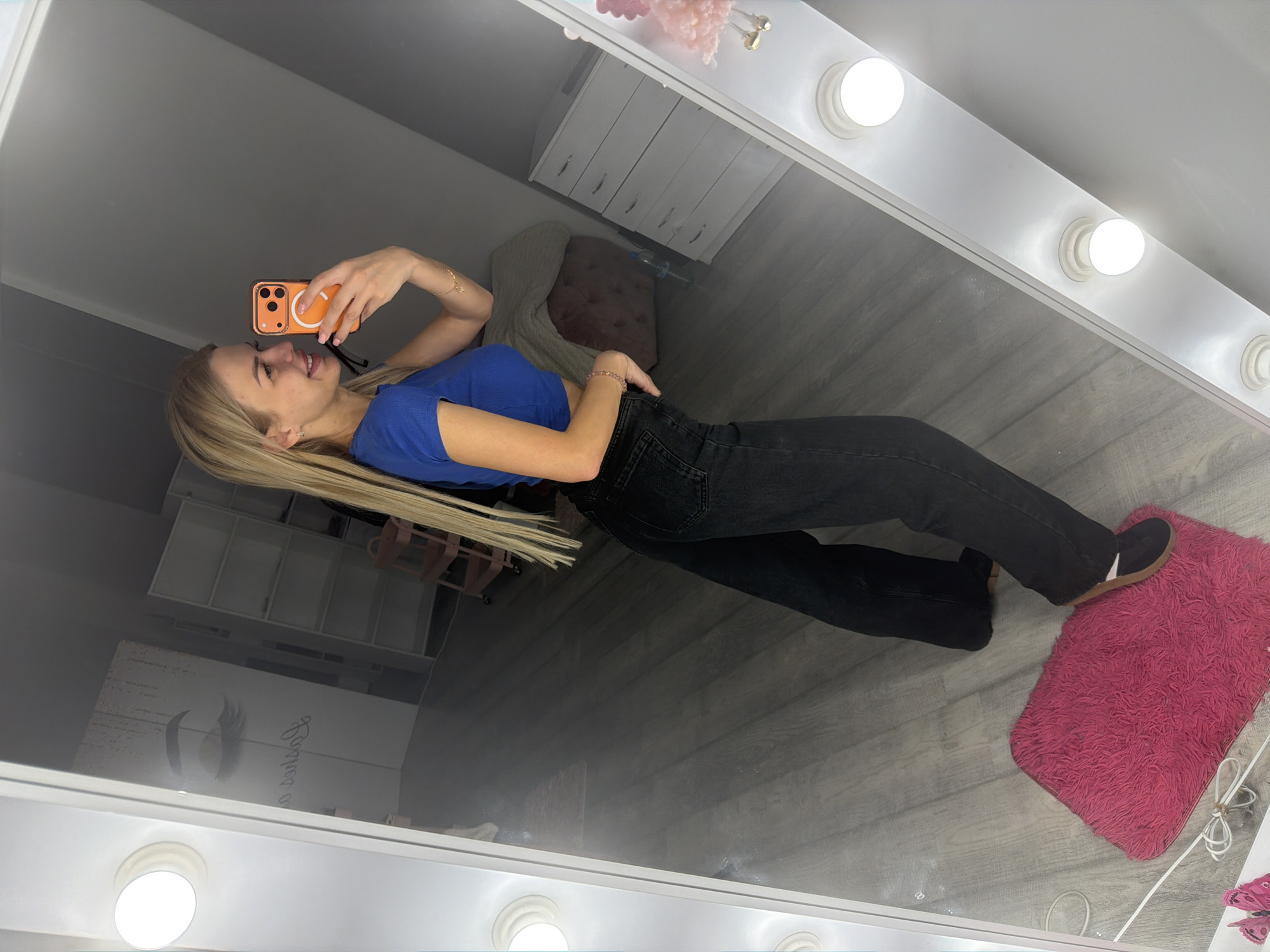 A blonde woman stands in front of a large illuminated mirror, taking a selfie with her orange-cased phone. She is wearing a blue crop top and black wide-leg jeans. Her right foot is on a bright pink shaggy rug. In the reflection of the mirror, a white chest of drawers, a pink stool, and shelves can be seen. The flooring appears to be grey wood laminate. The setting seems to be a room in Kyiv, Ukraine, possibly a beauty salon or dressing room, given the branding visible on a wall in the reflection that says "Lashes..." with an eyelash graphic.