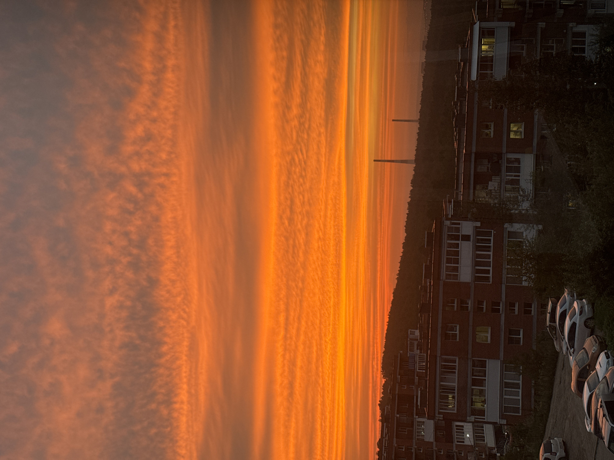 An aerial, medium shot captures a vibrant sunset over the city of Markova, Russia. The sky is a brilliant canvas of orange and red, with horizontal bands of clouds stretching across the horizon, creating a textured, layered effect. In the foreground, the tops of several apartment buildings are visible, characterized by their brick facades and numerous balconies. Some windows are lit, suggesting occupancy. A gentle, tree-covered slope rises behind the buildings. In the distance, two tall, slender industrial chimneys stand silhouetted against the fiery sky. The overall scene depicts a tranquil urban landscape bathed in the warm hues of sunset.