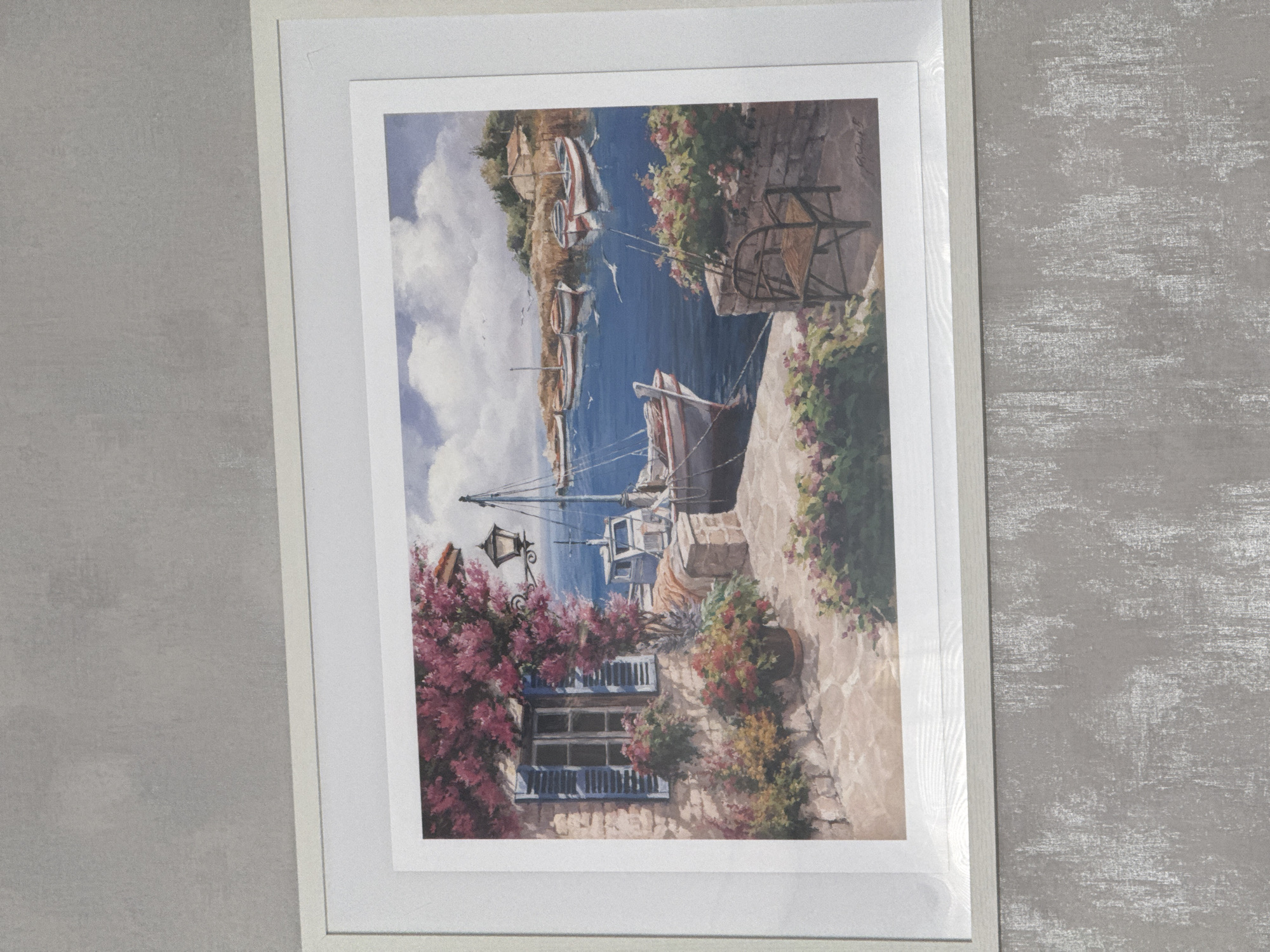 A framed painting depicts a seaside village scene. The painting features boats docked in a harbor, stone buildings with flower-filled window boxes, and a stone pathway. The artwork is framed with a white matte and a light gray frame. The background is a mottled gray wall. There is no information present in the image that can be used to identify the location as Lovech, Bulgaria.