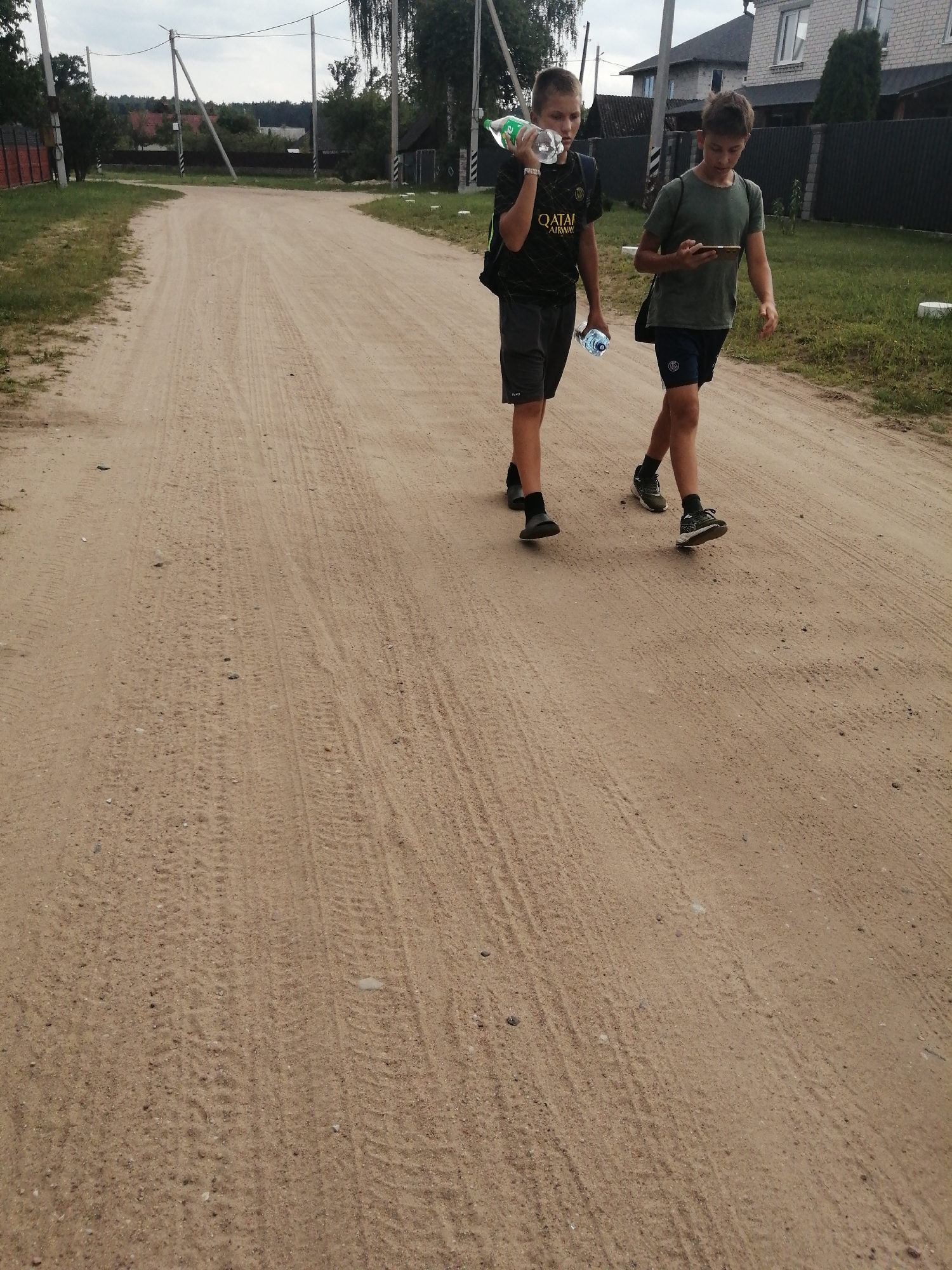 Two young boys are walking down a dirt road. The boy on the left is wearing a black t-shirt with "QATAR AIRWAYS" on it, grey shorts, and black shoes. He is holding a plastic bottle of water in his right hand and has a black backpack on his left shoulder. The boy on the right is wearing a green t-shirt, navy blue shorts, and black and white sneakers. He is looking at his phone, which he holds in his right hand, and has a black messenger bag over his left shoulder. The road is unpaved and appears to be a country road, with tire tracks visible in the dirt. In the background, there are houses, trees, and utility poles. The sky is overcast. This scene is consistent with a residential area in Slonim, Belarus, given the architectural style of the houses and the rural setting.