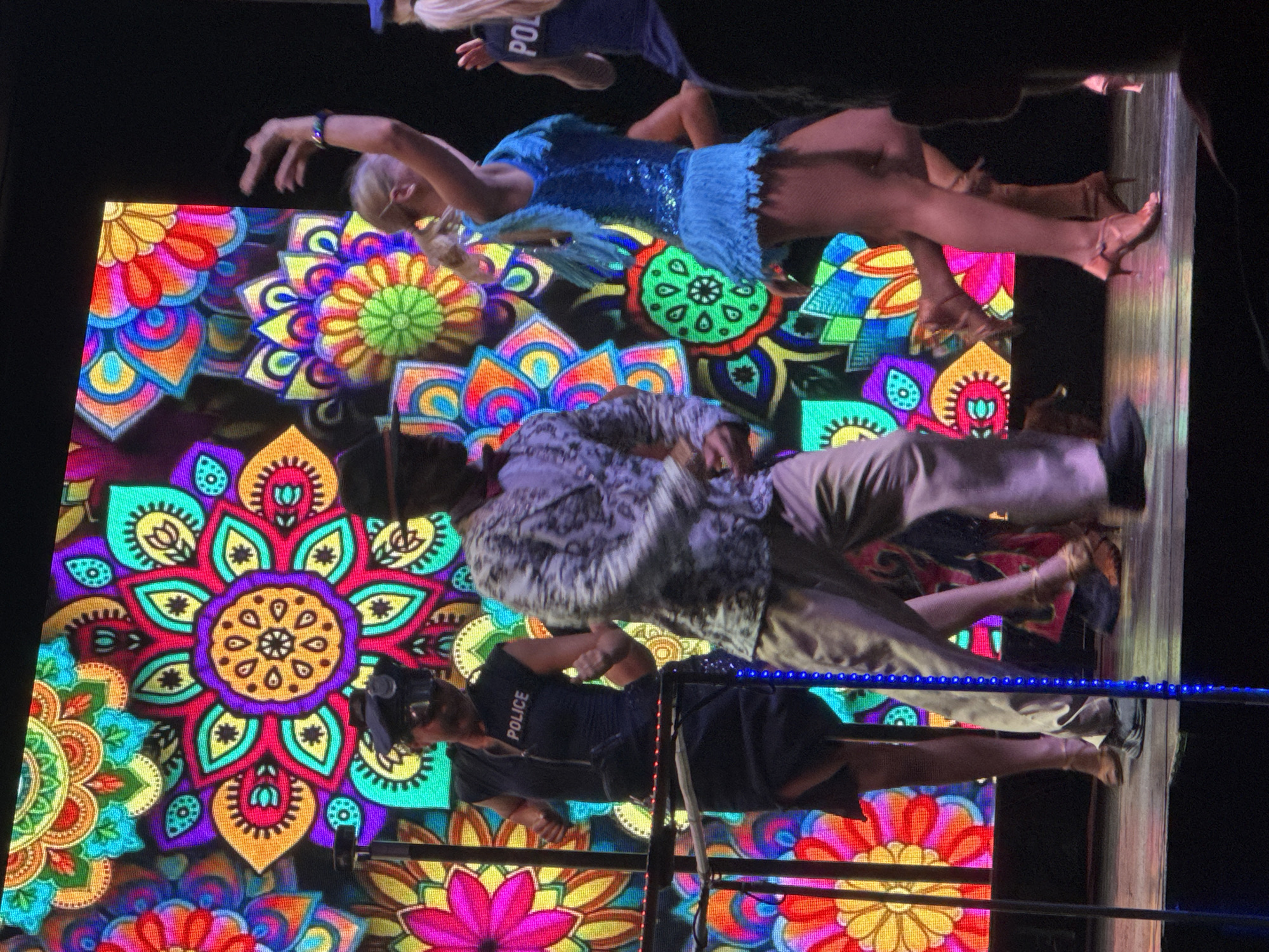 A group of people are performing on a stage in front of a large, brightly colored, mandala-patterned LED screen. The person in the foreground left appears to be dressed as a police officer, wearing a dark uniform with "POLICE" written on the chest and a police hat. To their right, a person in a patterned jacket and tan pants is dancing. On the right side of the stage, a person in a sparkling blue outfit with fringe is also dancing, with another person in a dark uniform with "POL" visible on the back to their right. The stage appears to be made of wood, and there are vertical poles with blue lights on the stage. The background image is abstract and floral, with vibrant colors like pink, orange, yellow, green, turquoise, and purple. The event is likely taking place indoors at a venue in Manavgat, Turkey, possibly a hotel or entertainment complex given the stage setup and performance.