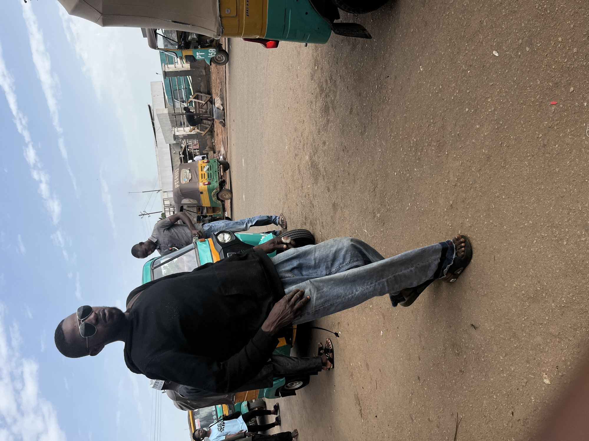 A man in sunglasses and a black hoodie walks across a dirt road in Gura Top, Nigeria. He wears blue jeans and black sandals. Several auto-rickshaws, a common mode of transport in Nigeria, are parked or driving on the road. The sky is cloudy. The scene appears to be a busy street market or transportation hub.
