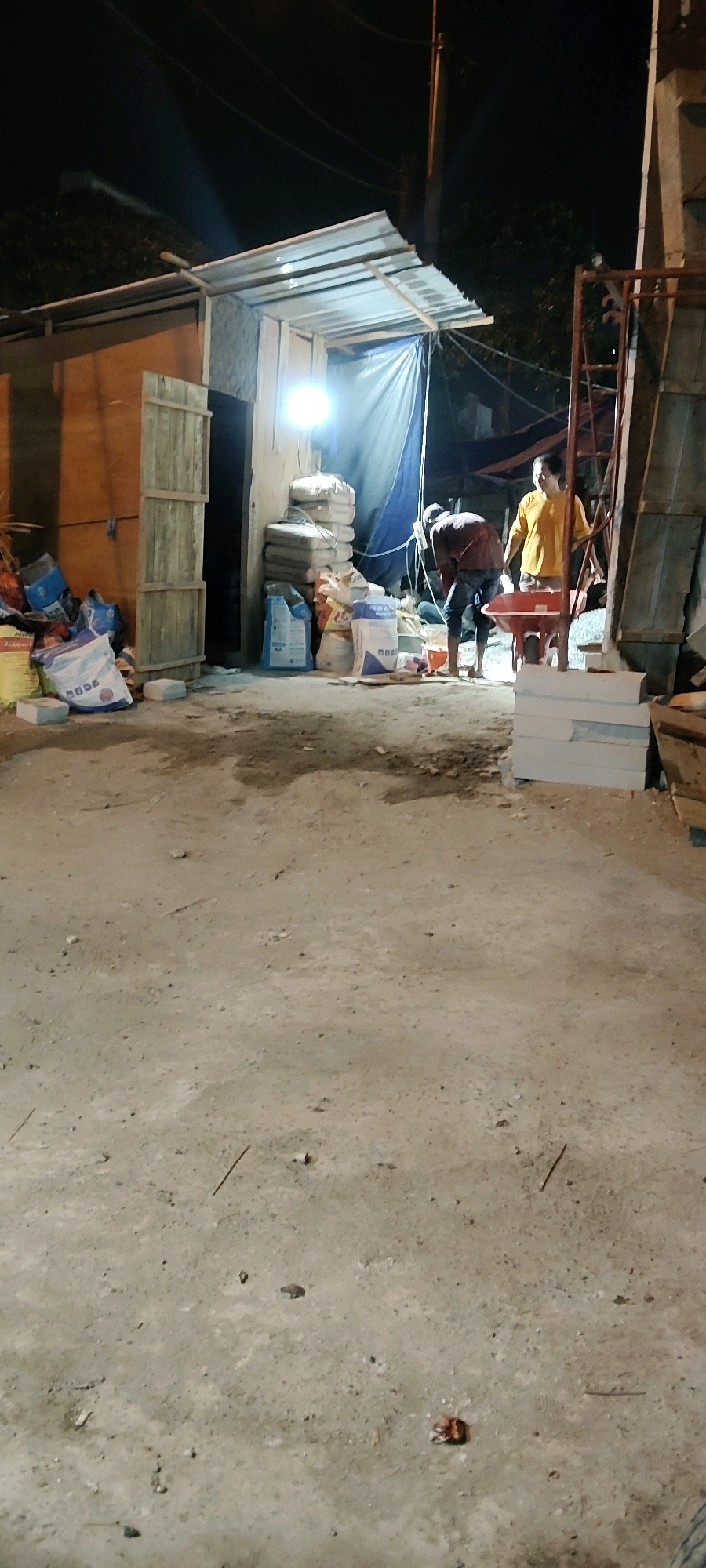 This image depicts a nighttime scene at a construction or material storage site in Cibatu, Indonesia. Two individuals are present. One, wearing a dark long-sleeved shirt and jeans, is bending over near a red wheelbarrow, appearing to be working or inspecting something. The other, wearing a bright yellow shirt, stands near the wheelbarrow, observing.
The environment includes a makeshift structure with a corrugated metal roof supported by wooden beams, covered by a blue tarp. Stacks of bags, likely containing cement or other building materials, are visible. Some of these bags are white with blue and red lettering, and one large yellow bag is also present. Wooden planks are leaned against a wooden structure on the left. In the foreground, the ground is composed of dirt and gravel, with scattered debris. Power lines are visible against the dark night sky above the structure. A ladder-like metal structure is to the right of the scene.