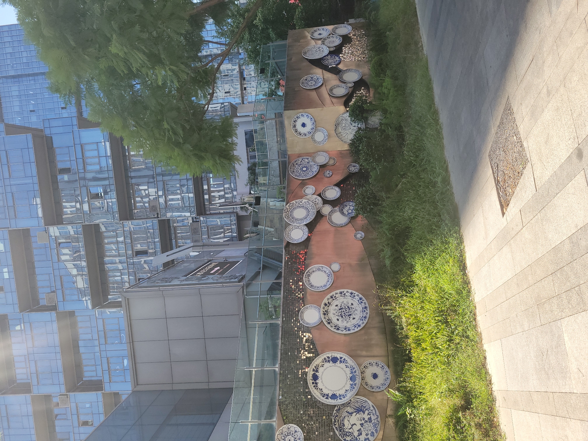 In Liangjiang, China, a wall is adorned with a collection of blue and white decorative plates. These plates, varying in size, are mounted on the wall's surface, creating an artistic display. The wall itself is textured and painted in earthy tones, with some sections appearing rough and others smoother. Behind the wall, lush green vegetation grows, including tall grass and ferns. In the background, a modern, multi-story building with a facade of glass and metal rises, reflecting the sky and surrounding environment. A large tree with dense green foliage is situated to the right of the building, partially obscuring it. The foreground features a paved walkway made of light-colored stone tiles, with a square grate embedded in the surface. The scene is illuminated by bright sunlight, casting shadows on the walkway.