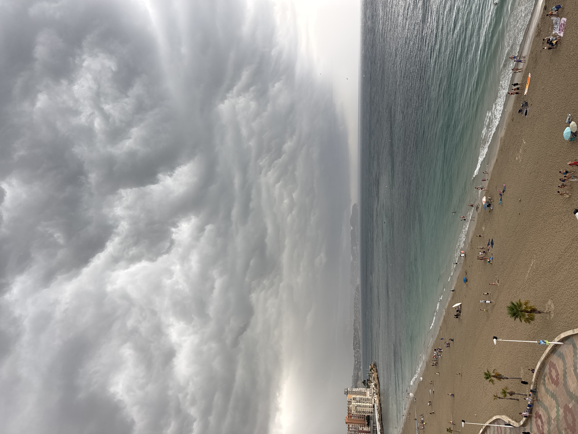 The image shows a wide view of a sandy beach in Calp, Spain, with a turbulent, cloudy sky overhead. The ocean is a deep blue and green, with moderate waves rolling onto the shore. The beach is populated with numerous people engaging in various activities: some are walking along the water's edge, others are sitting or lying on towels, and some are playing in the sand. There are palm trees scattered across the beach, and a few colorful umbrellas and beach chairs are visible. In the background, a town with tall buildings lines the coast, and a distant mountain range can be seen under the dramatic sky. The overall impression is of a beach scene under an impending storm.